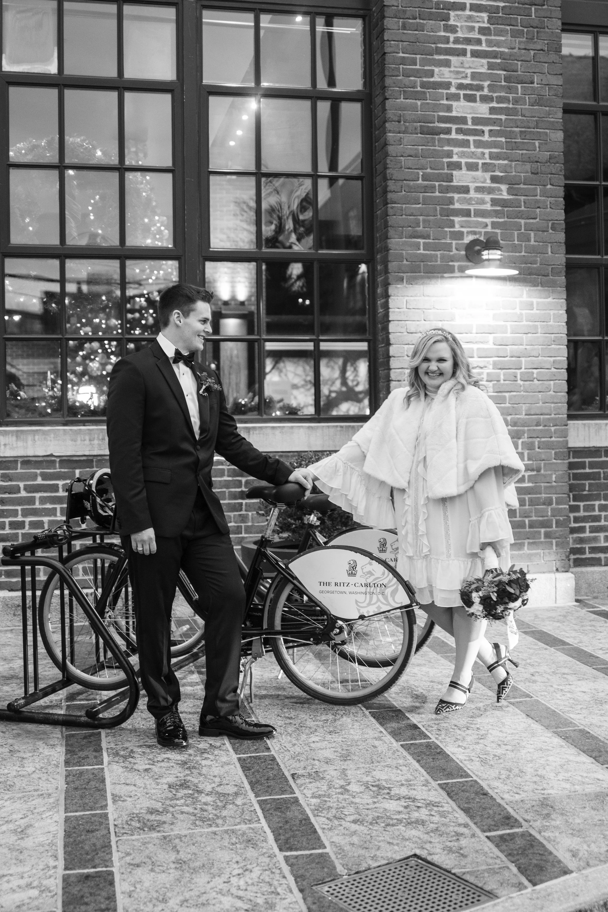A man and woman holding hands next to bicycles outside a brick building with large windows. The woman is smiling and holding a bouquet of flowers. The man is dressed in a tuxedo, and the woman is wearing a light-colored dress and heels. There is a Ch