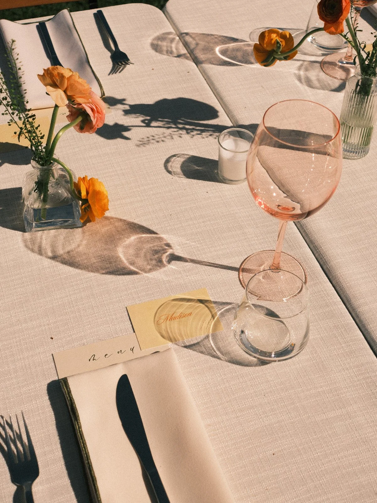 A table setting showing pink and clear glassware, small flower vases with orange and pink flowers, a candle, and place cards with elegant writing on a light-colored tablecloth, captured in sunlight.