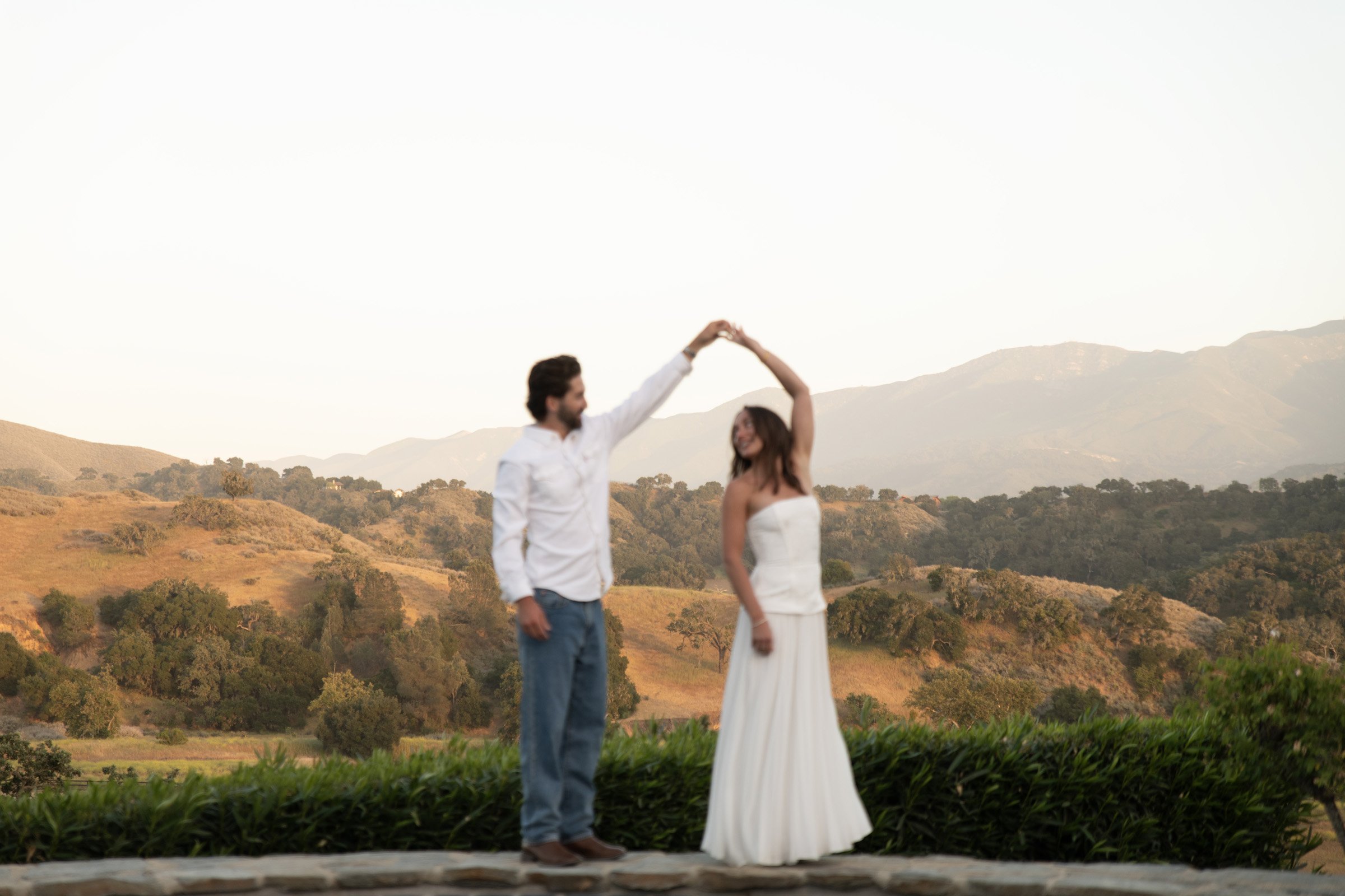 A couple in wedding attire dancing outdoors with rolling hills and mountains in the background, during sunset.