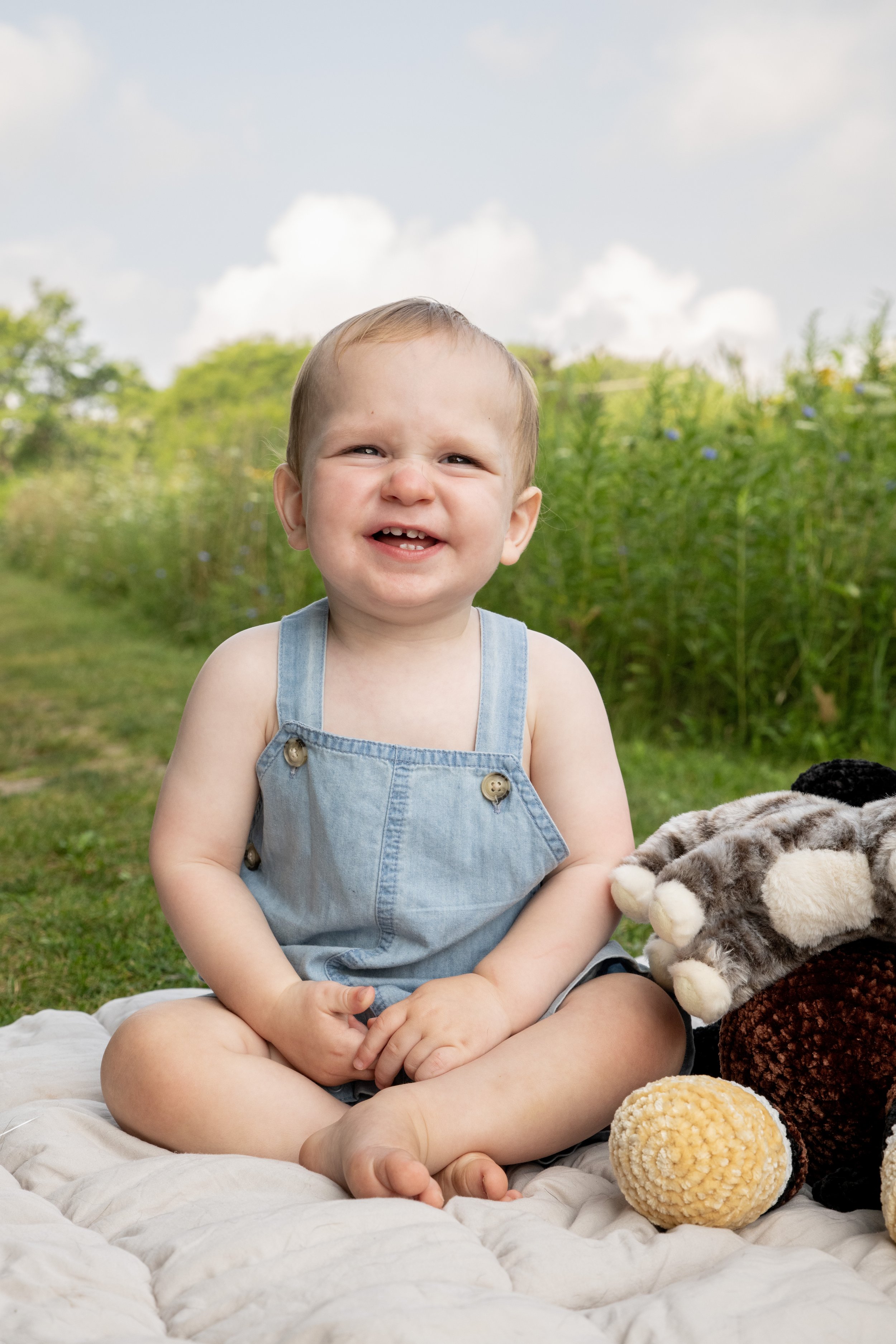 A smiling young child with short blonde hair, wearing denim overalls, sitting outdoors on a quilt with plush toys nearby, on a grassy field with green plants and trees in the background under a partly cloudy sky.