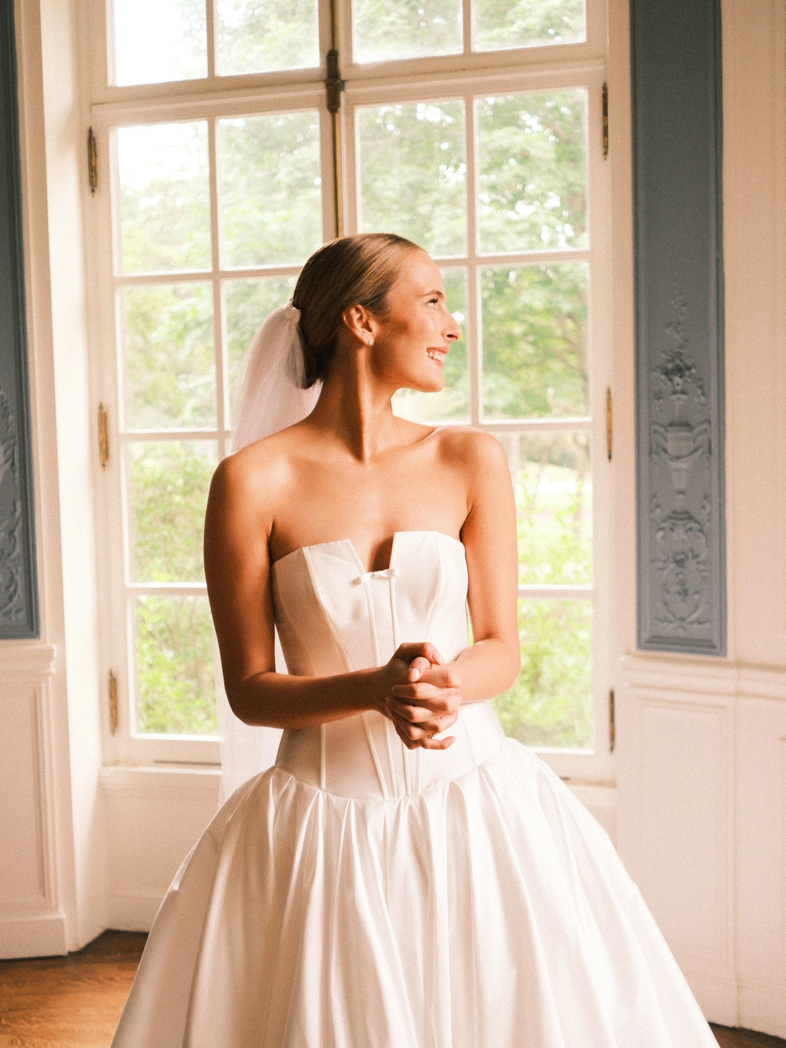 A smiling woman in a wedding dress with an updo hairstyle and veil, standing indoors near a window with greenery outside.