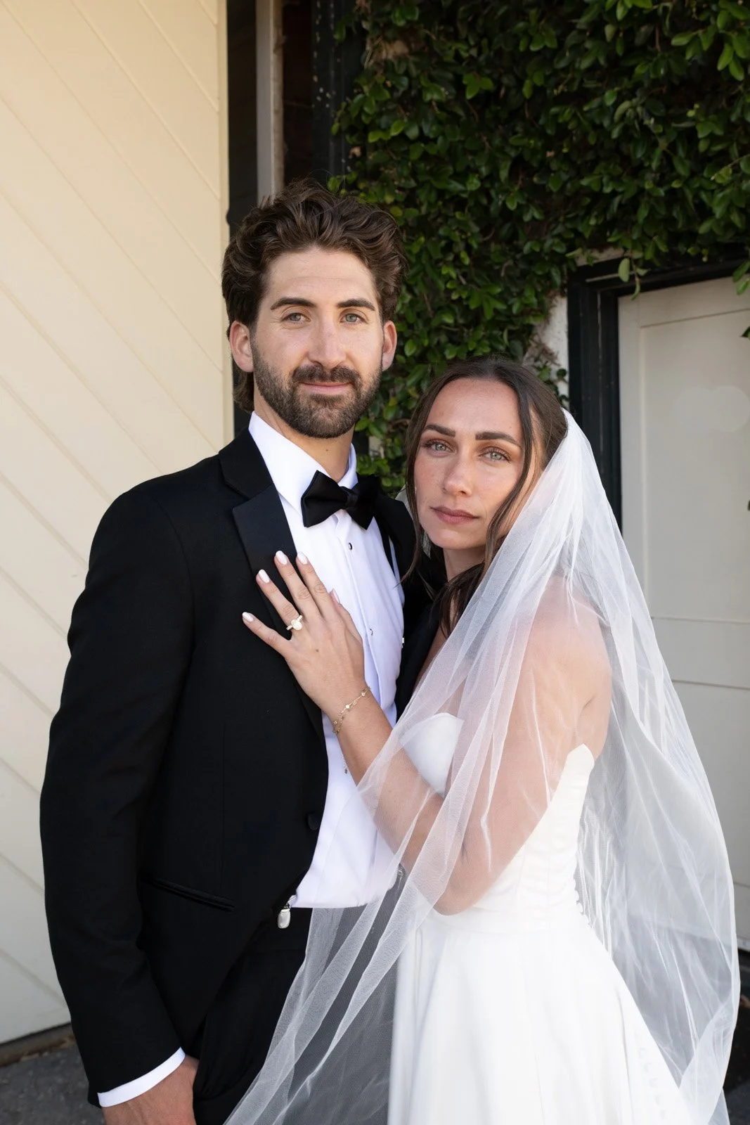 A newlywed couple posing outdoors, the groom with dark hair and beard in a black tuxedo, bowtie, and white shirt, and the bride with long dark hair and a veil dressed in a white wedding gown, standing close together with the bride's hand on the groom