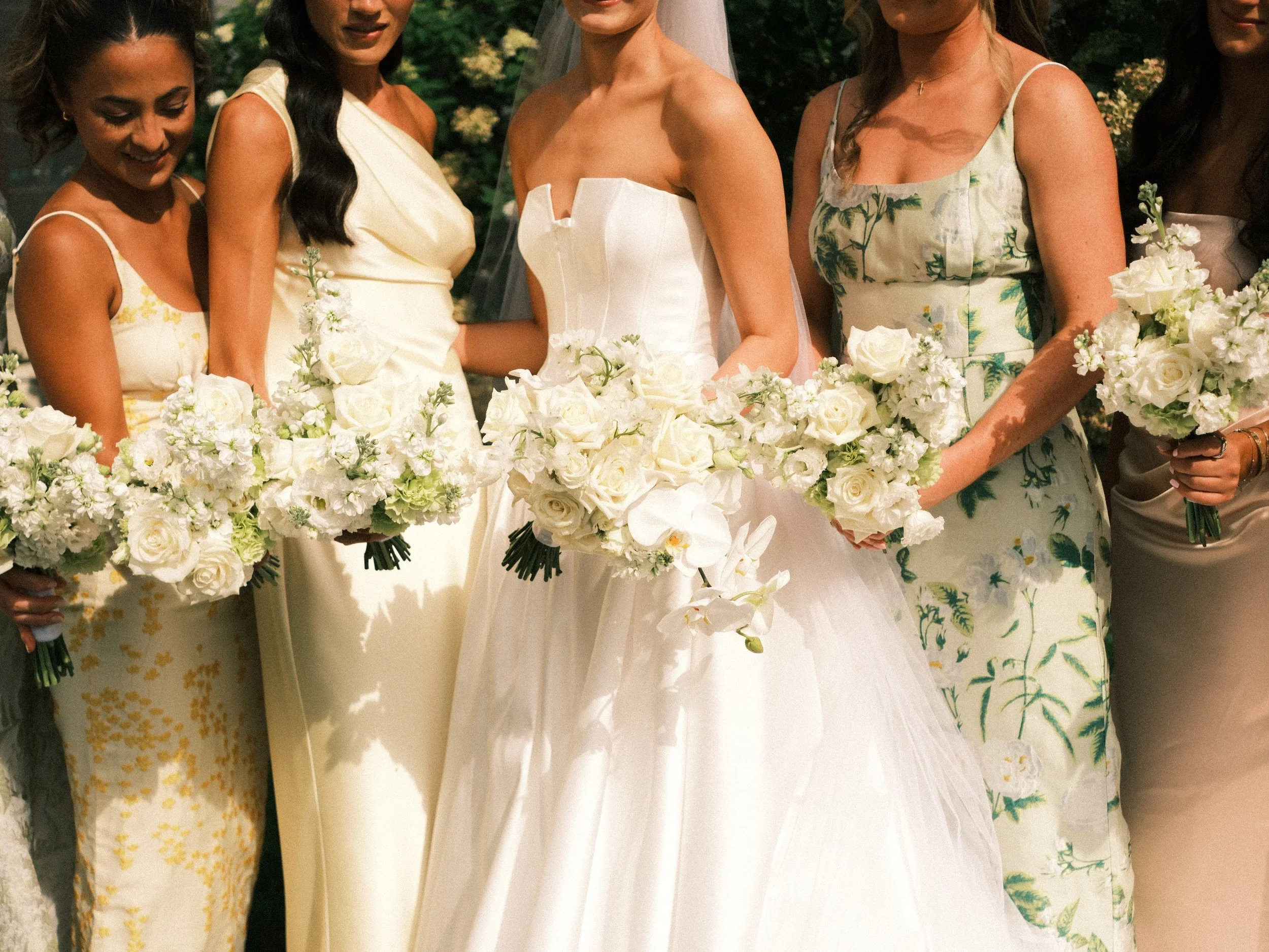A woman in a white wedding dress standing among women holding white bouquets, outdoors with greenery in the background.