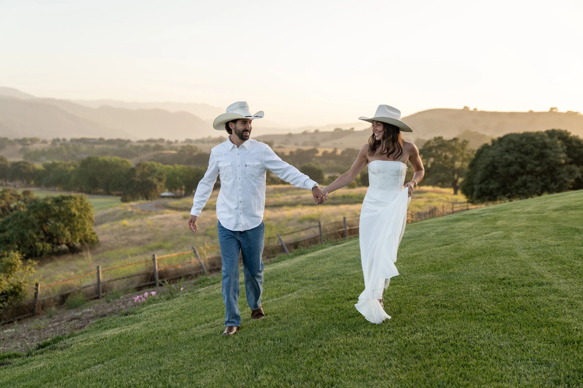 A man and woman dressed in Western attire, holding hands and smiling while walking on grass in a scenic outdoor setting with hills and trees at sunset.