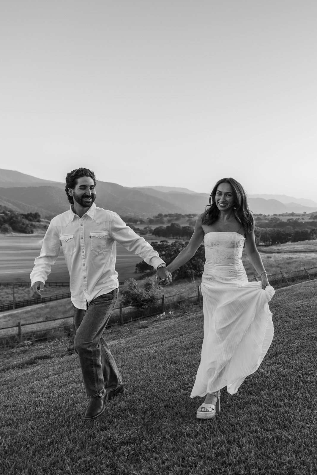 A black and white photo of a smiling couple holding hands and running on a grassy hill with a scenic landscape of rolling hills and trees in the background.