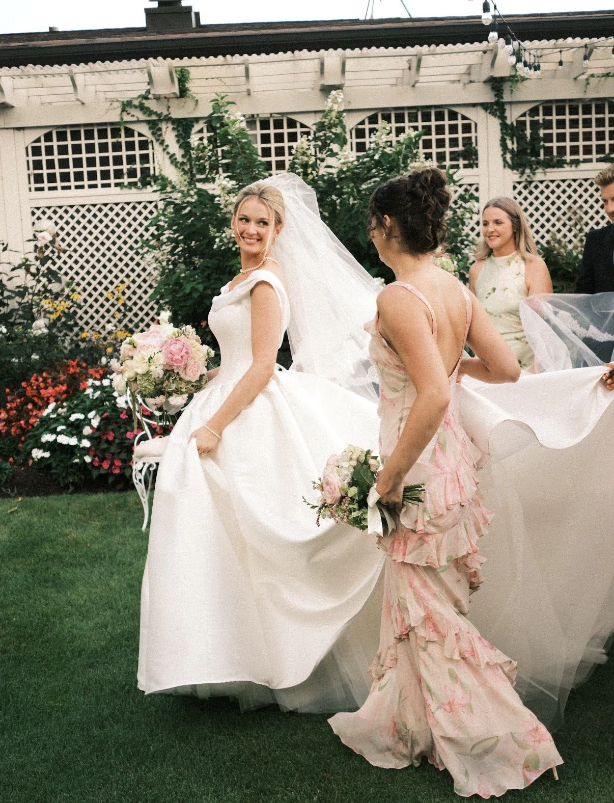 A bride in a white wedding dress holding a bouquet of pink and white flowers, smiling, while wedding guests surround her in a garden with colorful flowers and a white lattice fence.