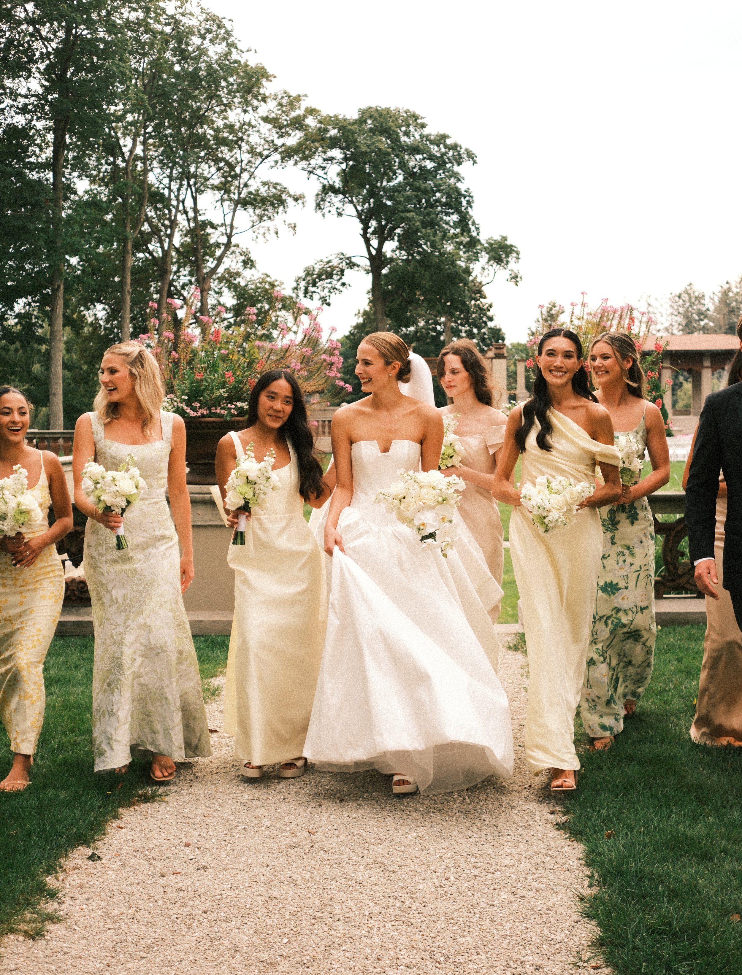 A bride in a white wedding gown walking outdoors with a group of bridesmaids in pastel dresses, holding bouquets, on a gravel path with grass and trees in the background.