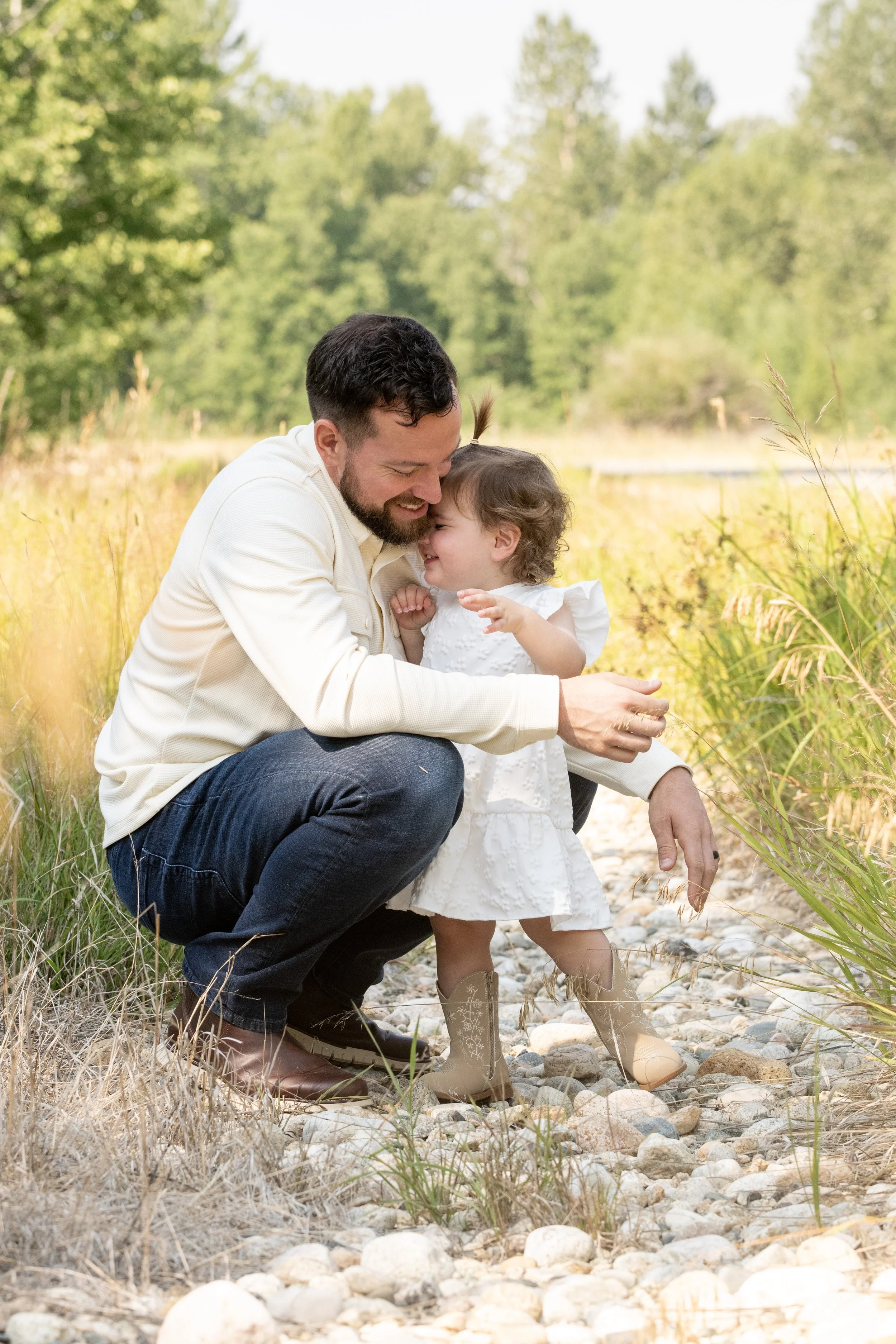 A man and a young girl are outdoors near a grassy area and rocks, sharing a joyful moment and smiling close together.