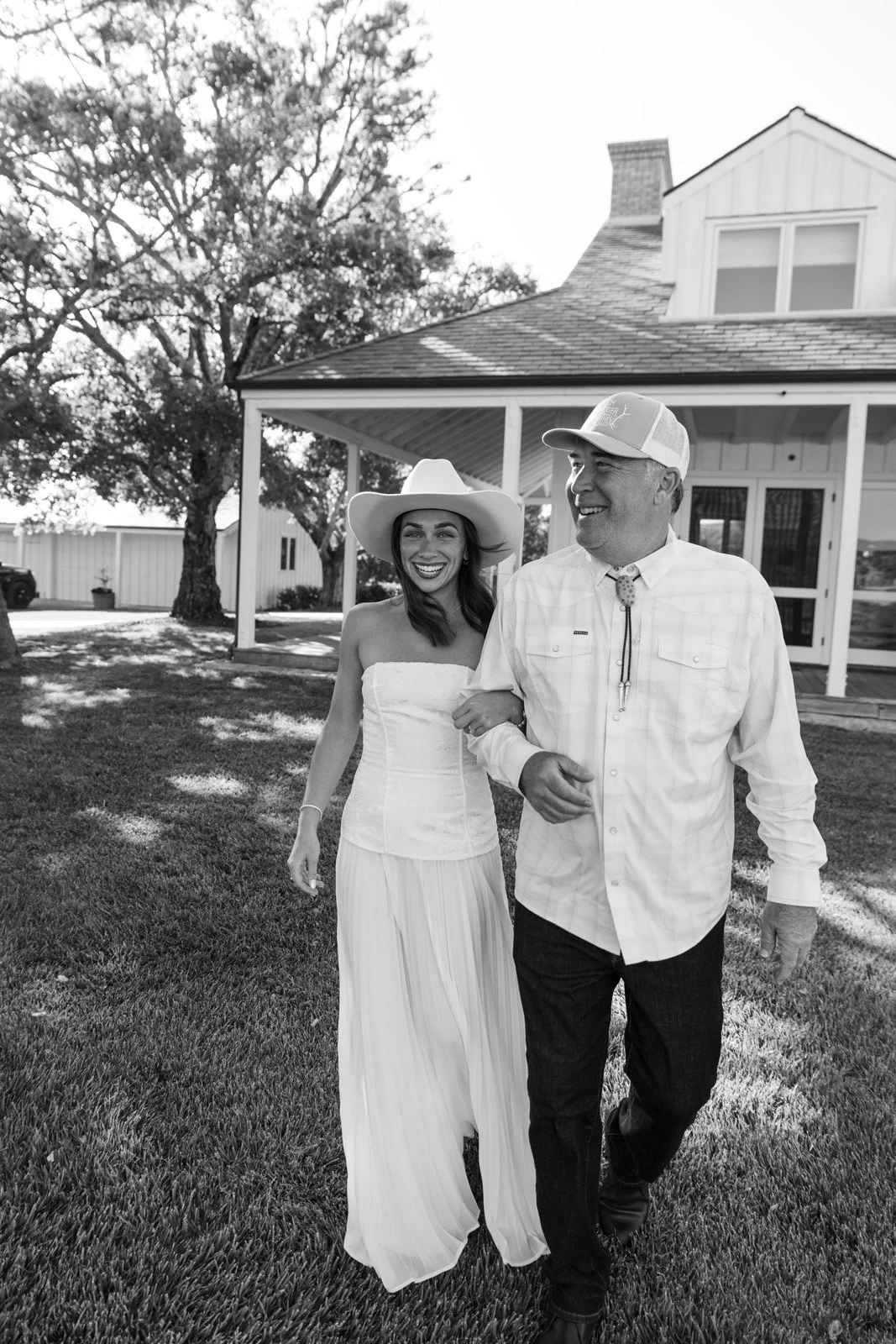 A woman in a strapless white dress and a wide-brimmed hat, and a man in a cowboy hat and checkered shirt, smiling and walking arm-in-arm outdoors in front of a house.