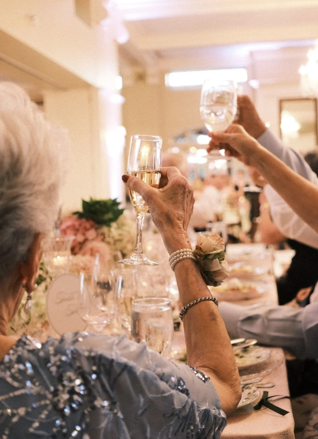Elderly woman with pearl bracelet and corsage raising a glass of champagne at a formal celebration or wedding dinner, with other guests holding glasses in the background.