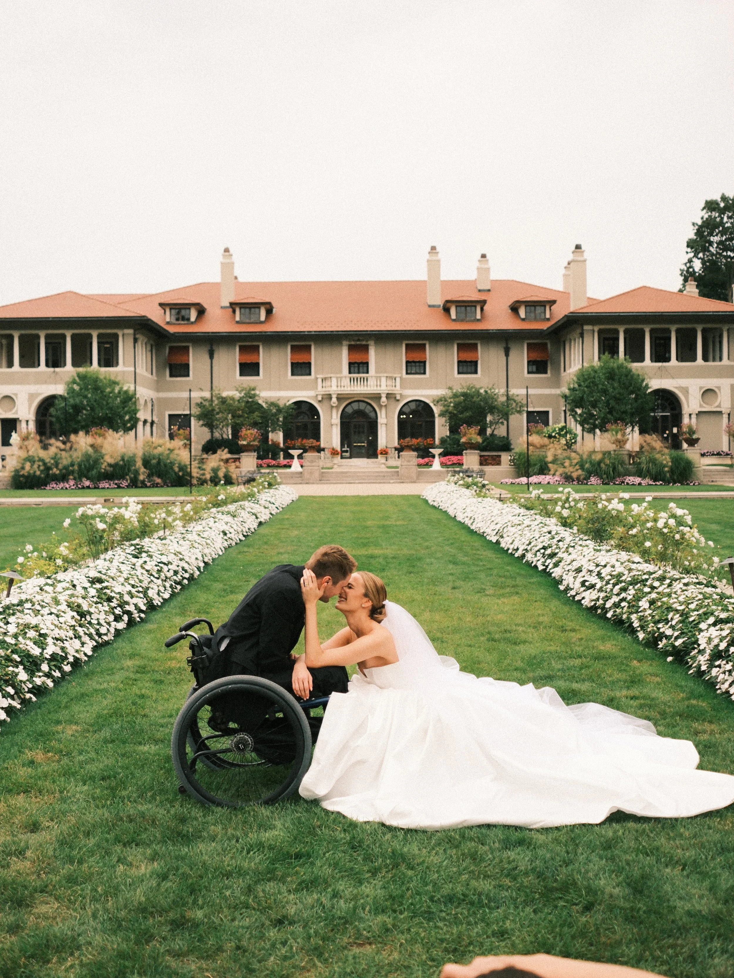 A bride and groom sharing a kiss outdoors on a wedding day. The groom is in a wheelchair, and the bride is wearing a white wedding gown. They are on a lawn with flower beds and a large, elegant building with a red roof in the background.