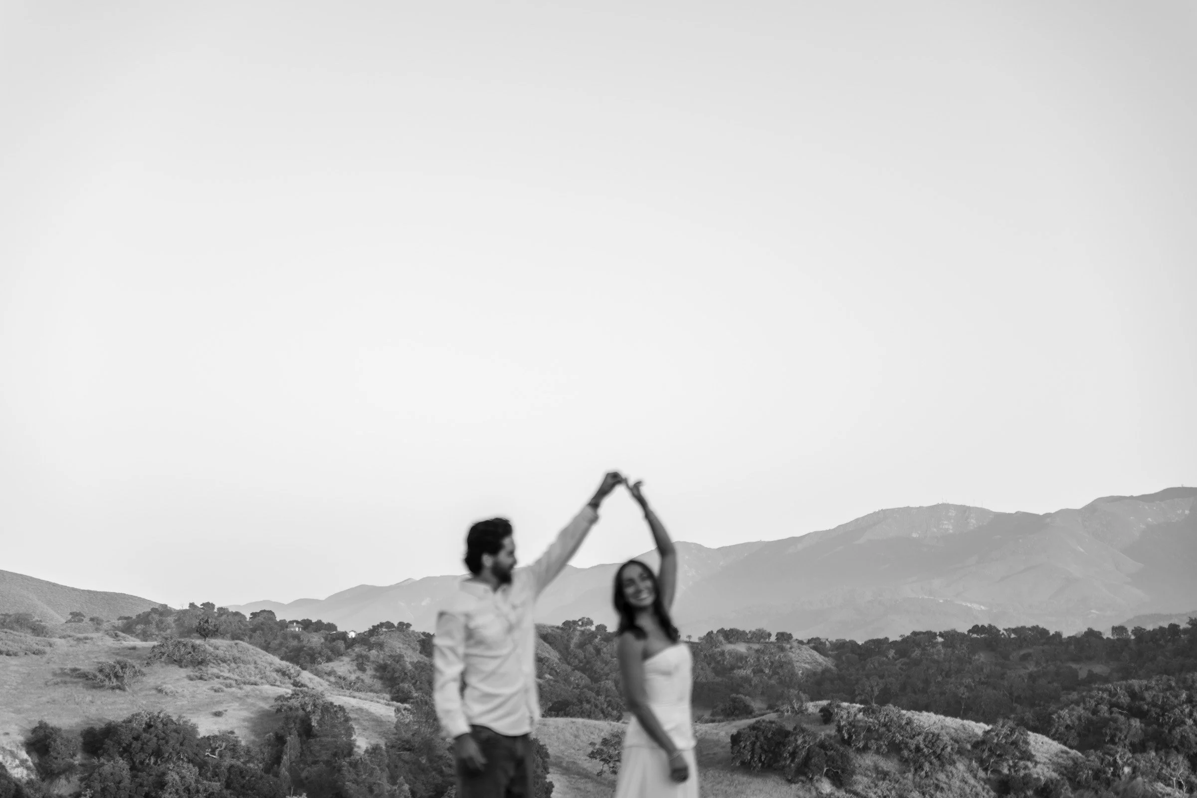 A black-and-white photo of a man and a woman standing outdoors in a scenic landscape with mountains in the background, holding hands above their heads in a triangle formation.