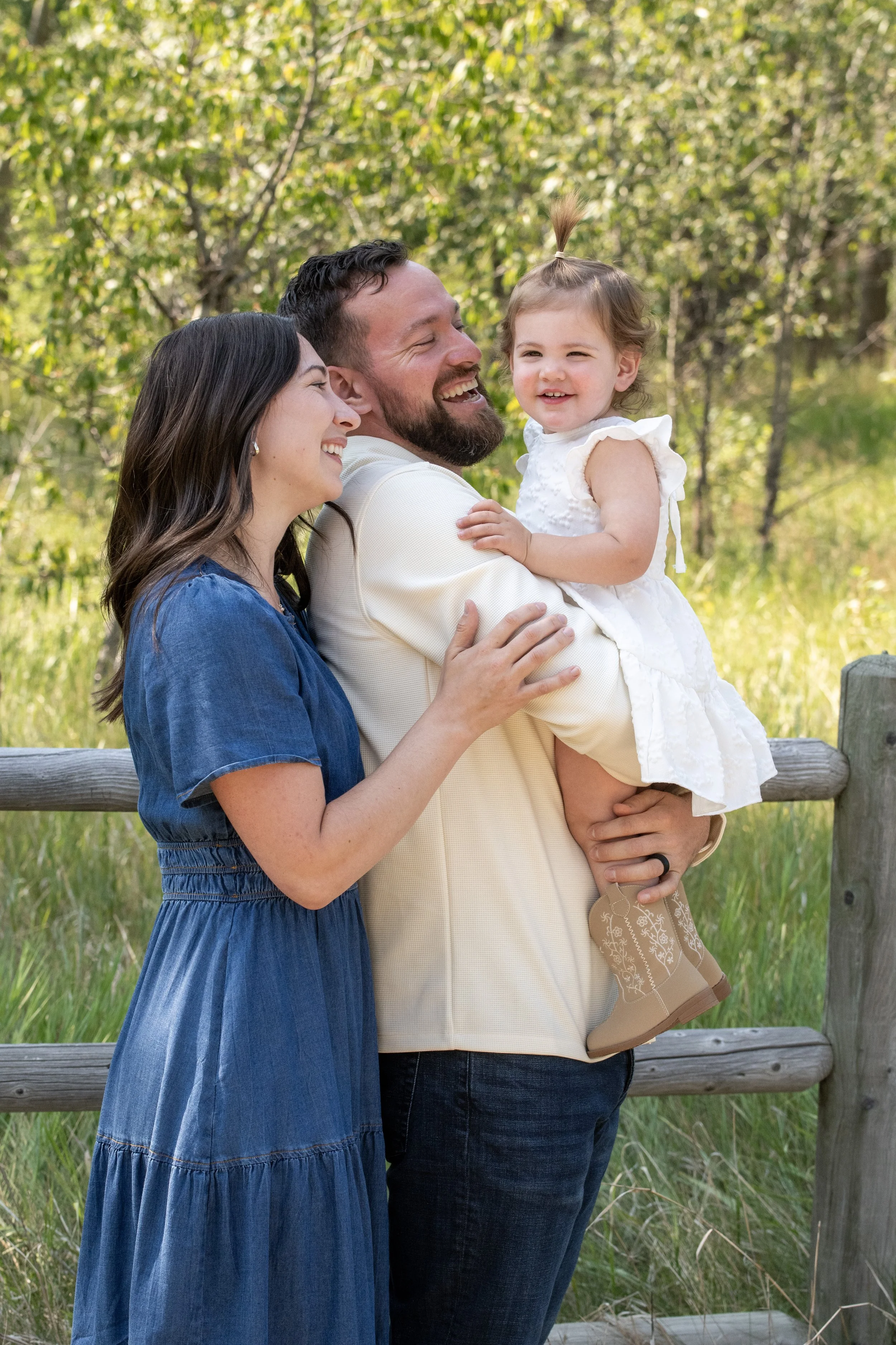 A happy family of three, a man, a woman, and a young girl, outdoors in a park with greenery, smiling and embracing each other.