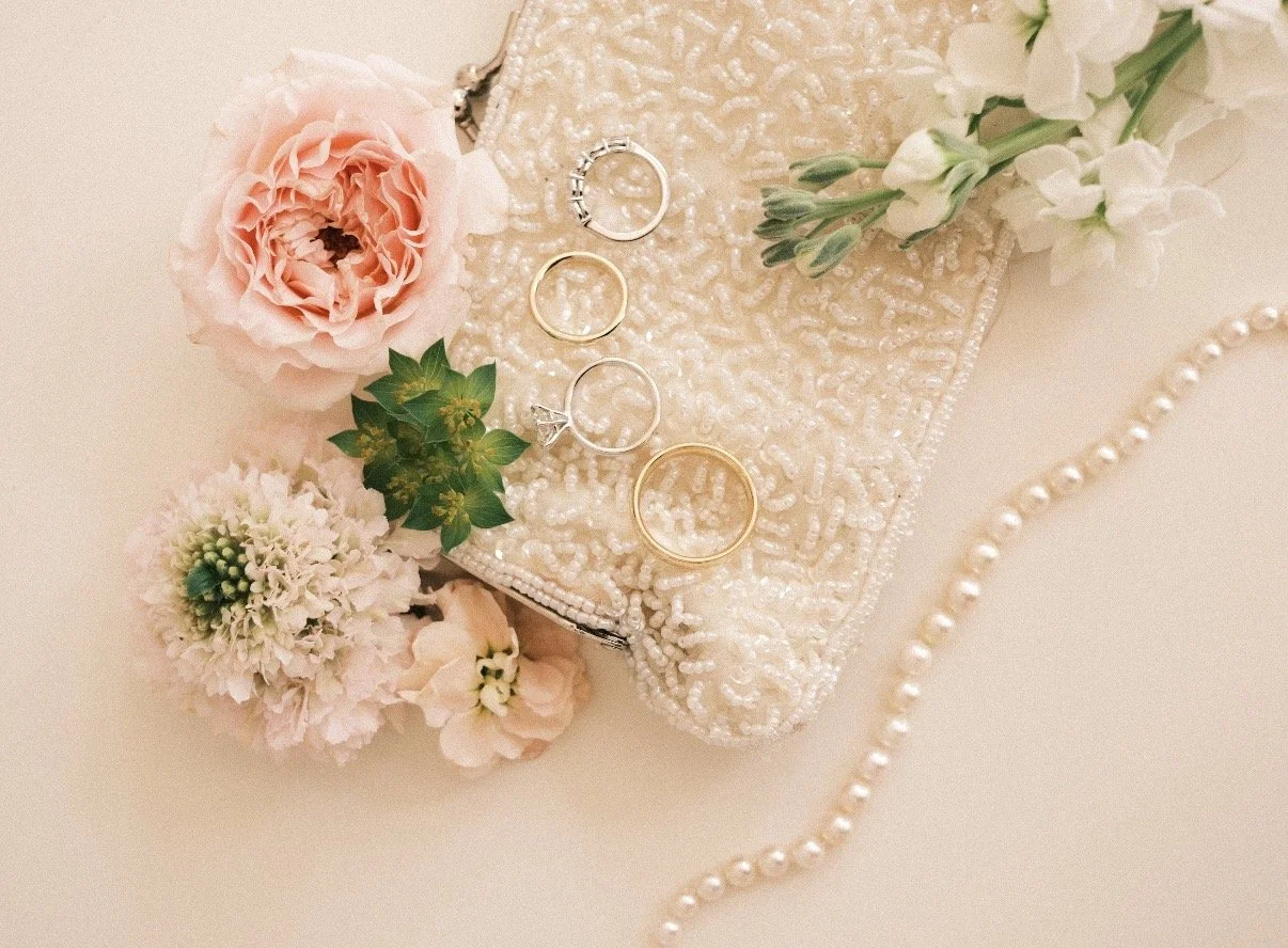 Close-up of a beaded clutch purse with pearl embellishments, adorned with wedding rings, surrounded by pink and white flowers, and a string of pearls.