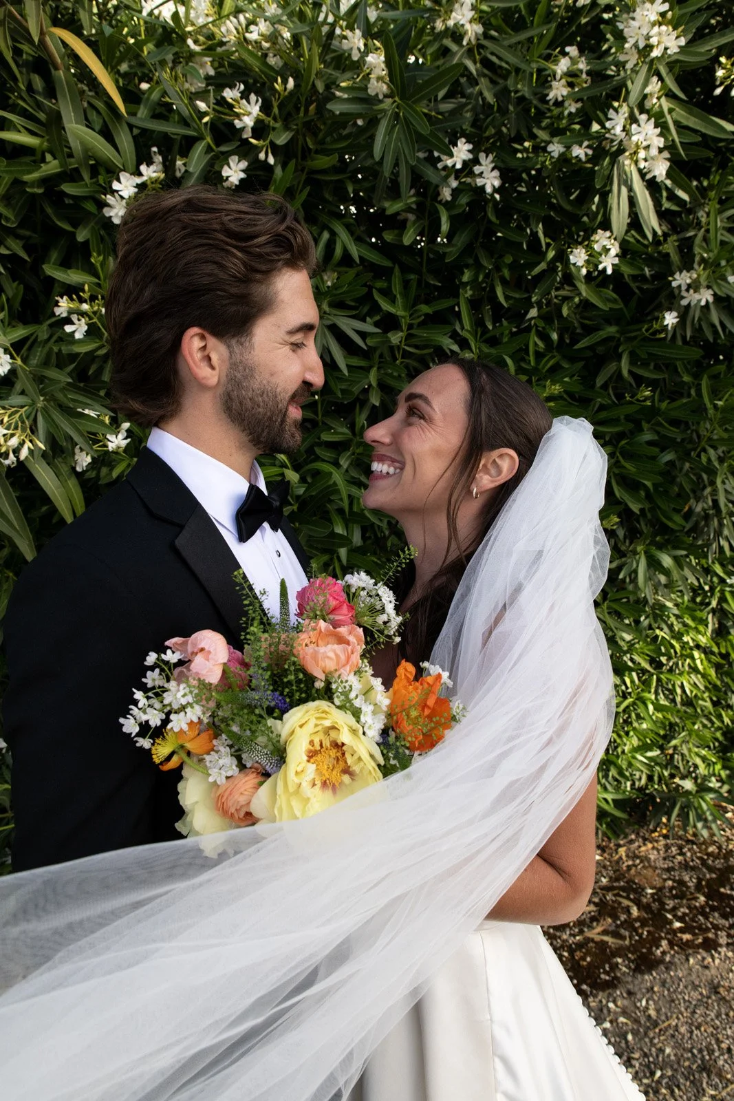 A bride and groom are smiling at each other, standing in front of green foliage with white flowers. The bride is holding a colorful bouquet of flowers and has a white veil.