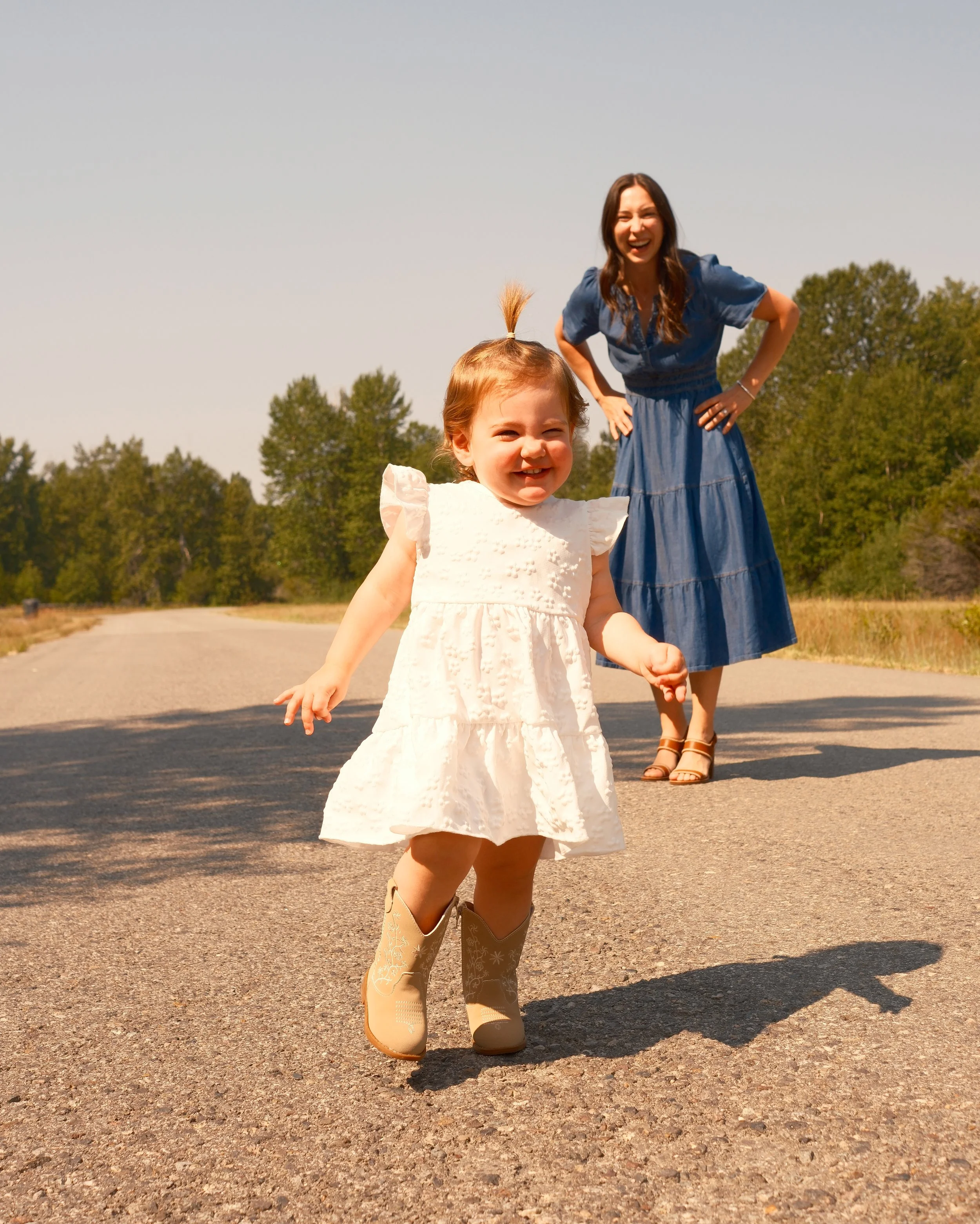 A young girl in a white dress and cowboy boots standing outdoors on a paved road, with a woman in a blue dress and cowboy boots standing behind her, smiling and laughing. They are in a rural setting with trees in the background on a sunny day.