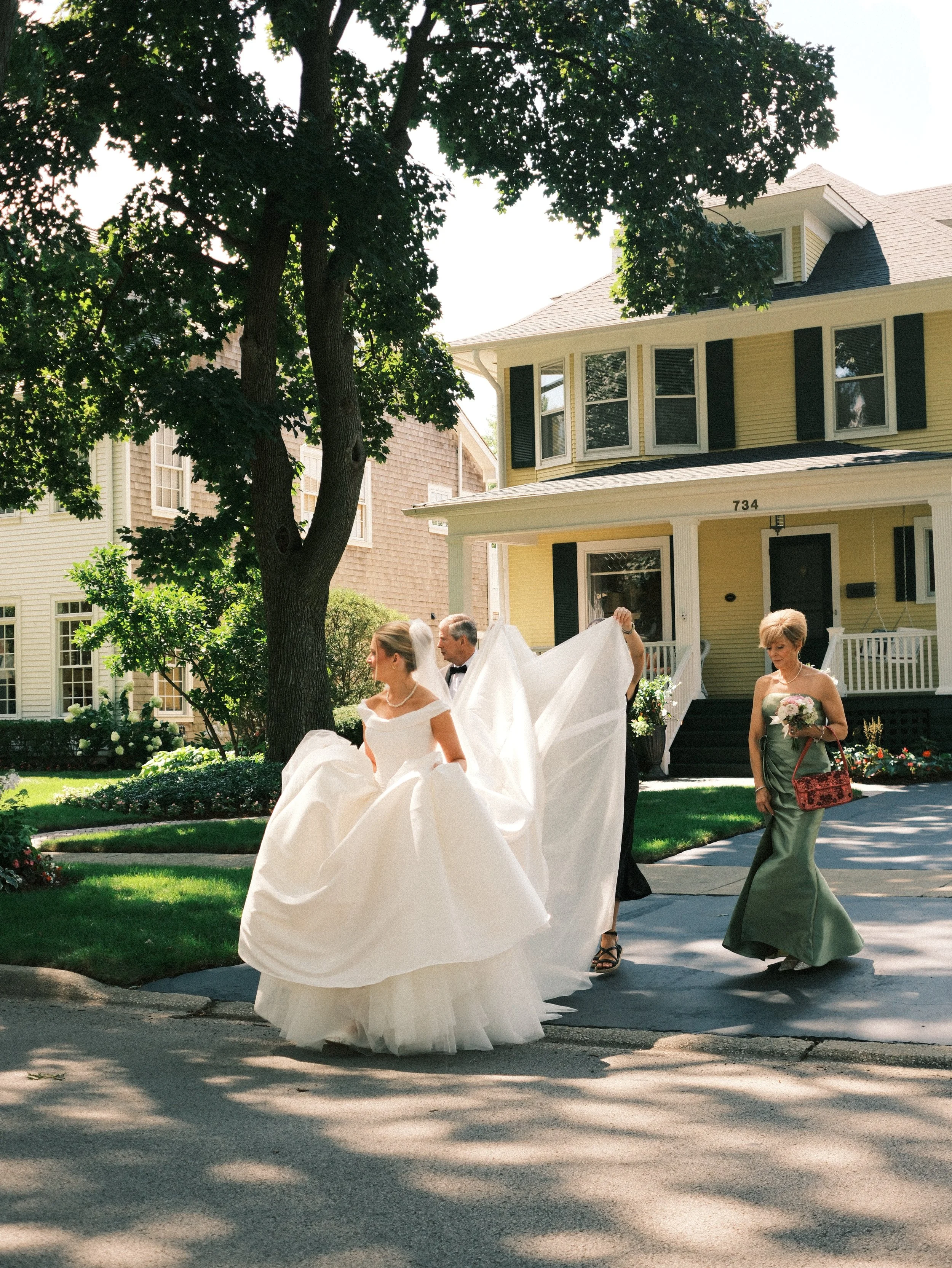 A bride in a white wedding gown walking on the sidewalk in front of a yellow house with black shutters, accompanied by women and a man, with some holding her train. The scene is shaded by a large tree.