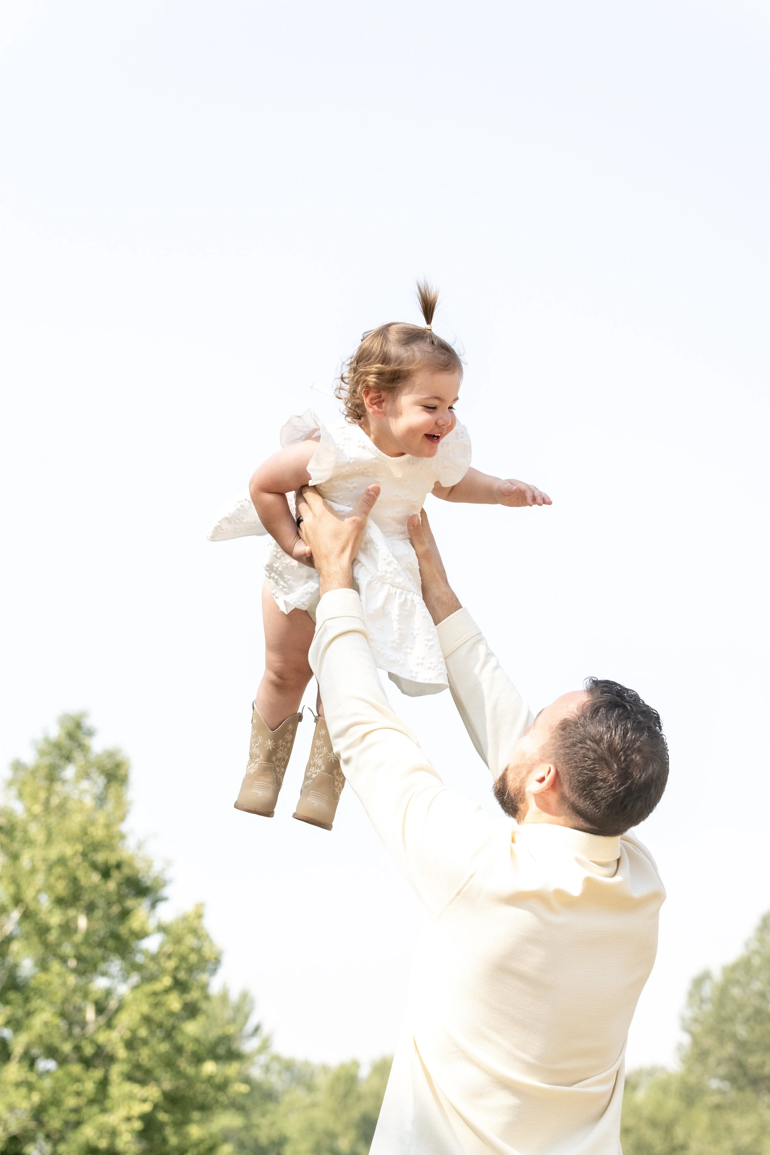 A man lifting a young girl into the air outdoors on a sunny day, with trees in the background.