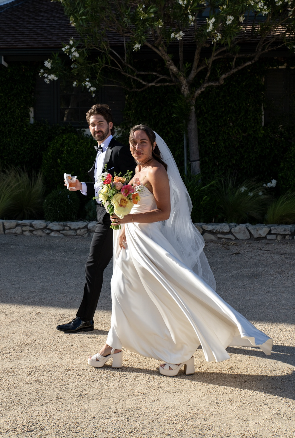 A bride and groom walking outside during their wedding, with the bride holding a colorful bouquet of flowers and wearing a long white wedding dress with platform heels, the groom in a black tuxedo with a bow tie, and a background of greenery and a tree.
