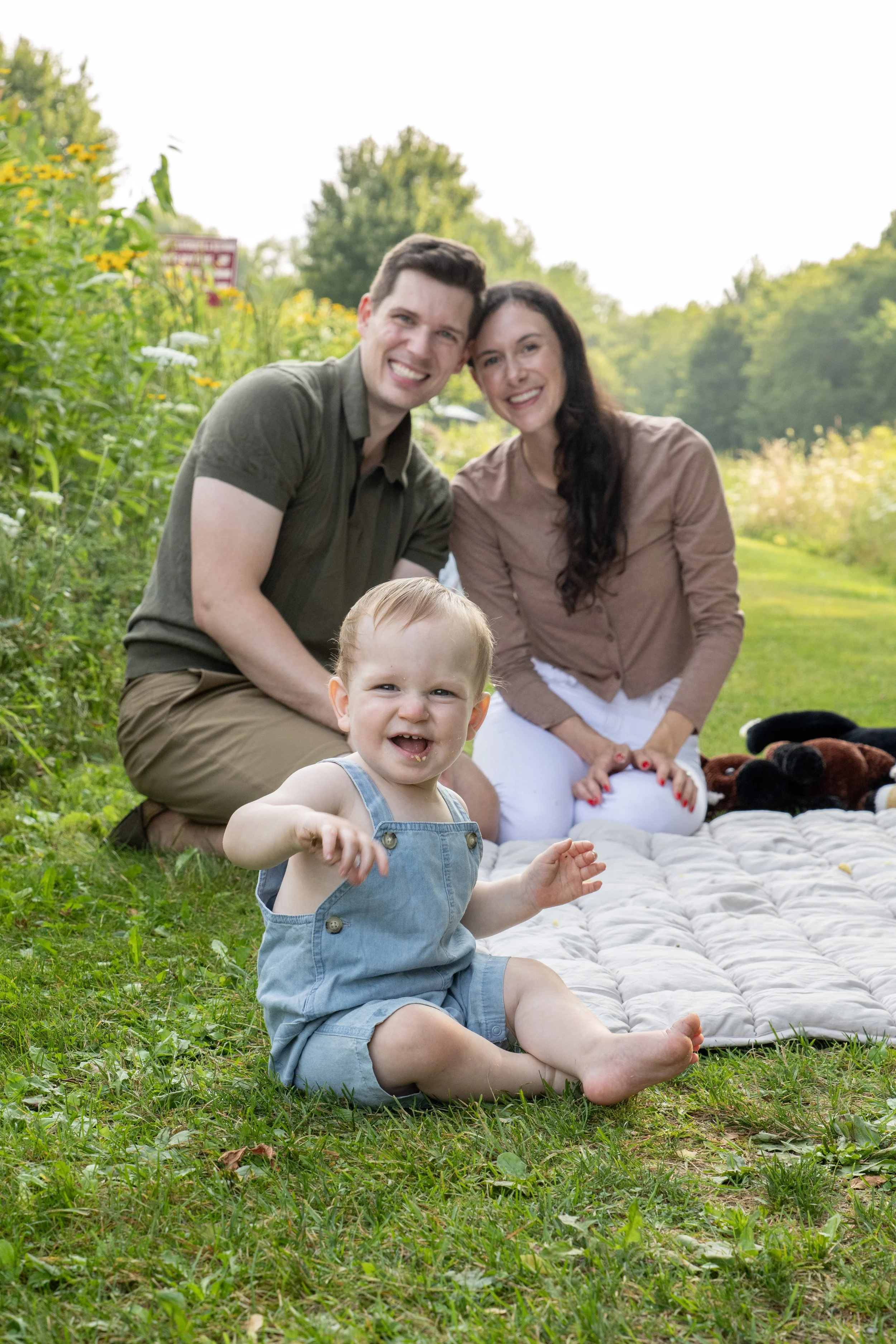 A happy family of three, including a young child, outdoors on a grassy field with a blanket and toy animals, surrounded by trees and colorful flowers, enjoying a sunny day.