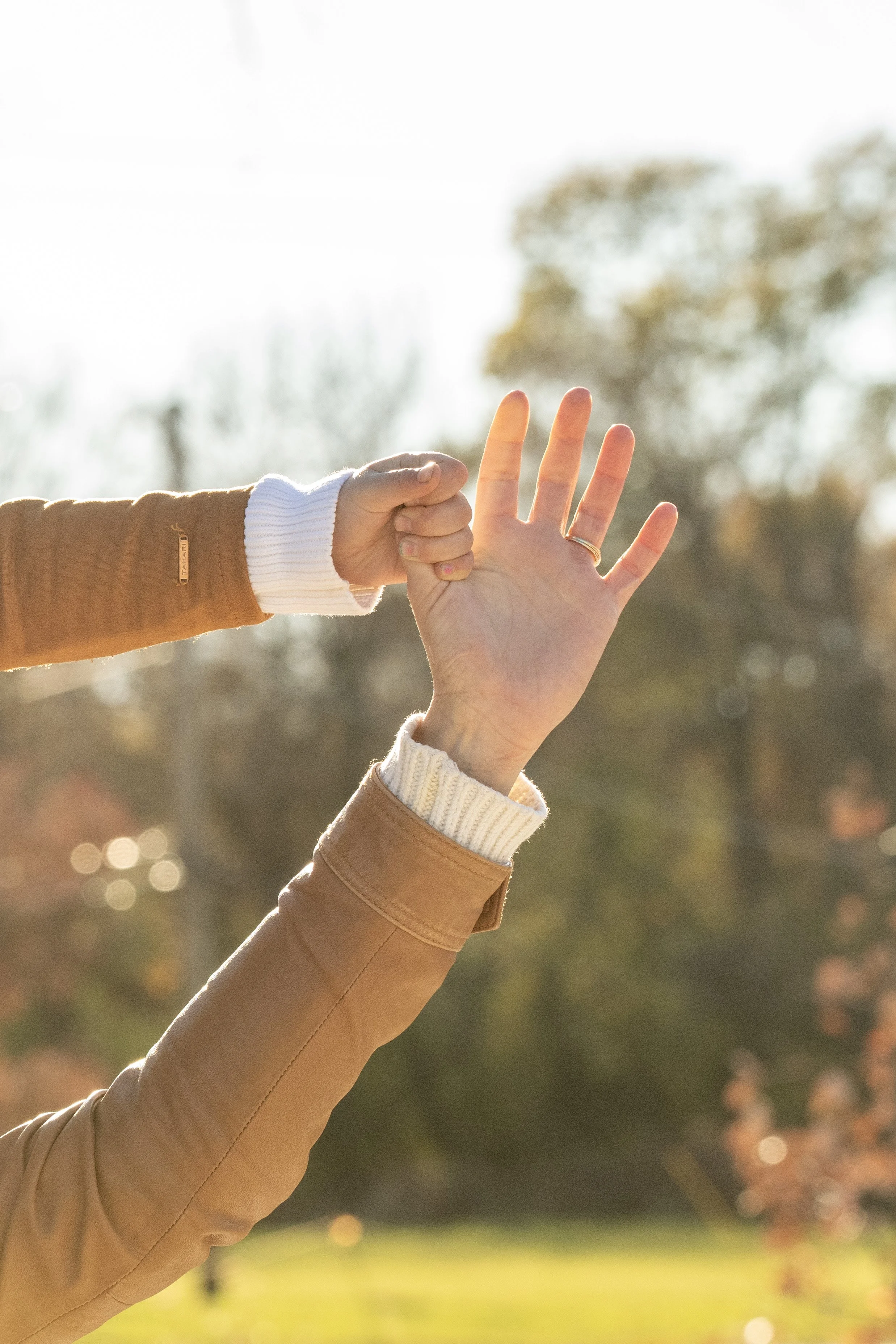A person wearing a tan coat and white sweater is holding up a hand with fingers spread in front of another person's hand, which is making a fist. The background features a blurred outdoor scene with trees and sunlight, suggesting fall or winter.