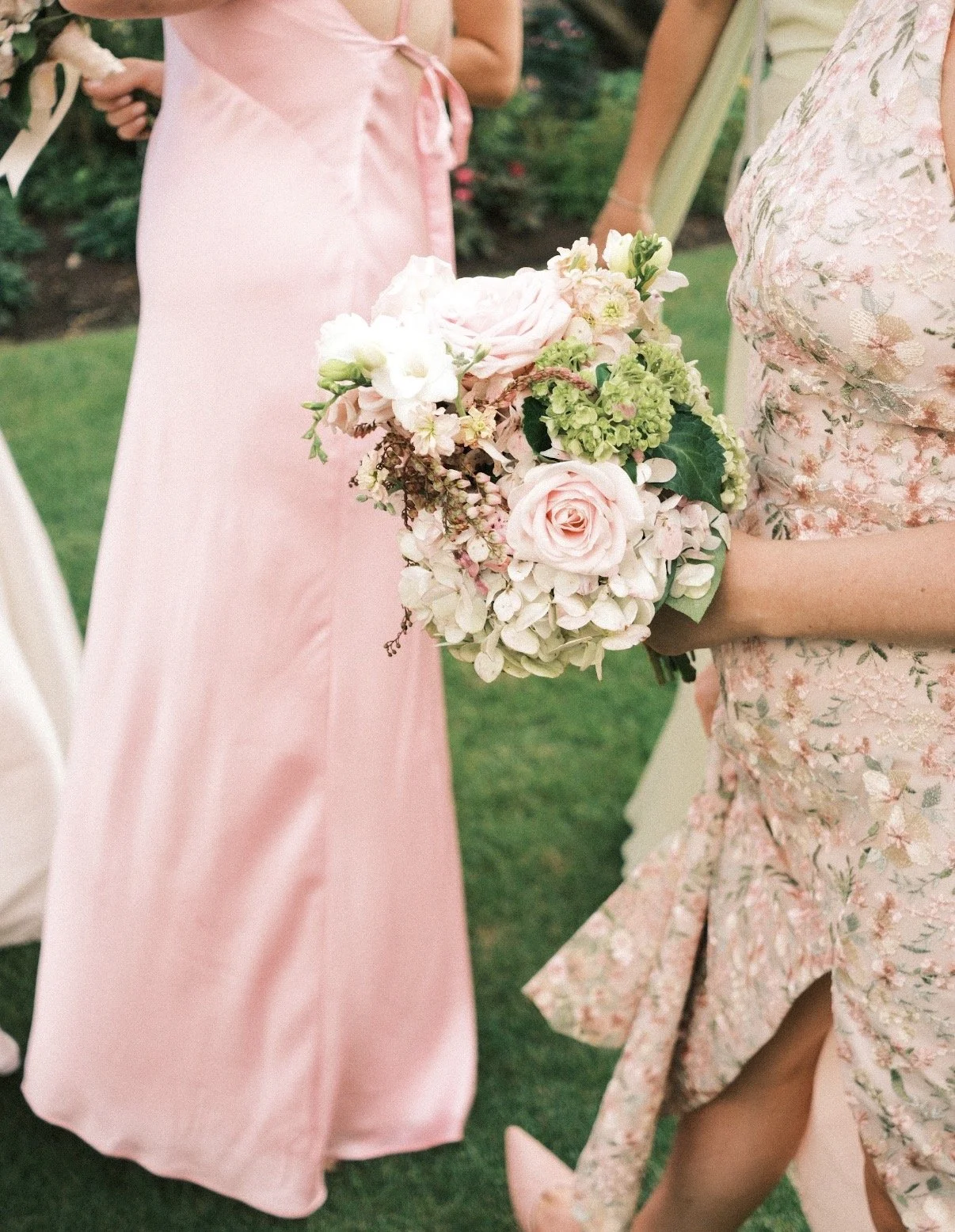 A woman holding a bouquet of pink and white flowers, with other women dressed in pink and floral dresses standing nearby, outdoors in a garden setting.
