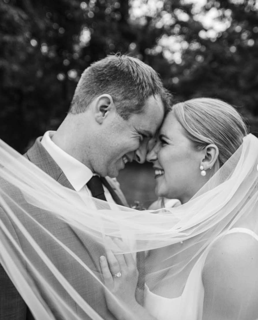 Black and white photo of a newlywed couple smiling with foreheads and noses touching, her veil draped over their faces, outdoors with blurred trees in the background.
