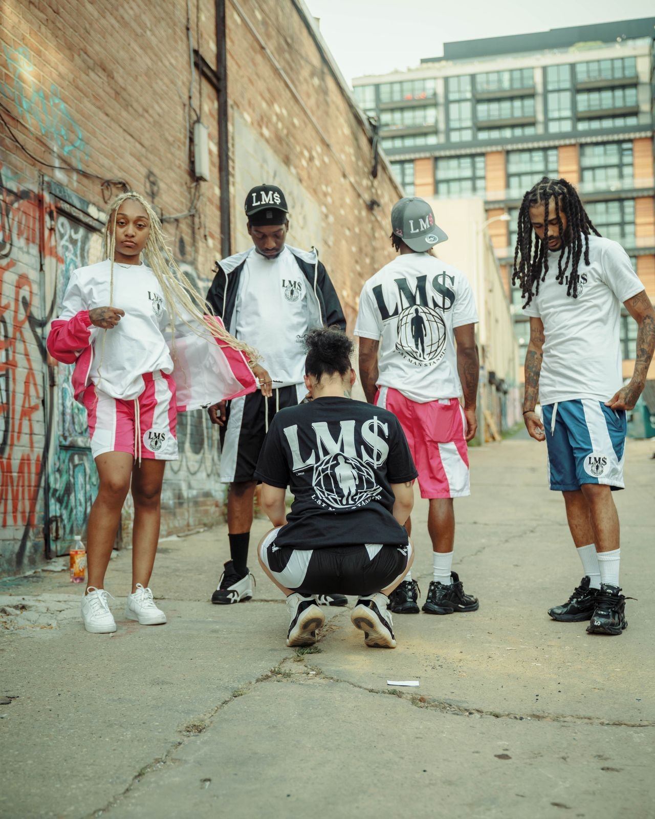 Group of five young people wearing matching streetwear outfits with 'LMS' branding, standing in an urban alleyway with graffiti-covered walls and tall buildings in the background.