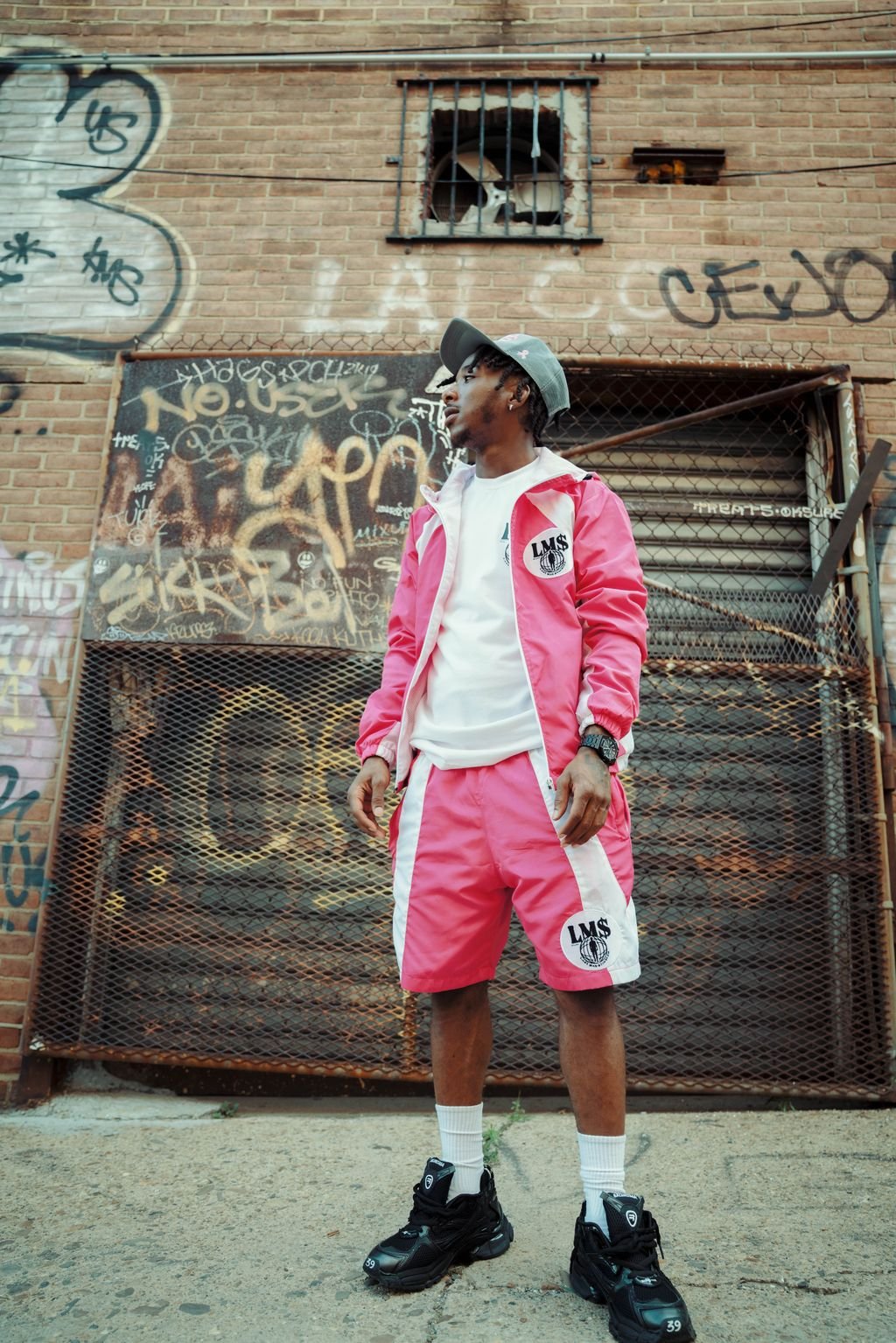 A young man wearing a pink and white tracksuit, matching shorts, a white t-shirt, and black sneakers with white socks, standing outdoors in front of a graffitied wall with old metal and brick structures.