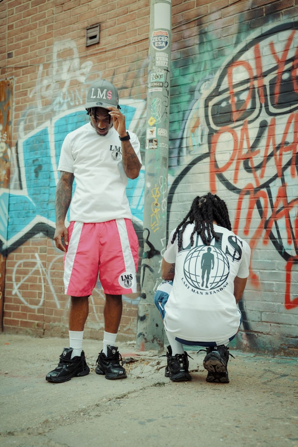 Two young men in streetwear stand against a graffiti-covered brick wall and metal pole in an urban setting. One man, wearing a gray cap and pink shorts with a white T-shirt, is looking down and adjusting his cap. The other man, with long dreadlocks,
