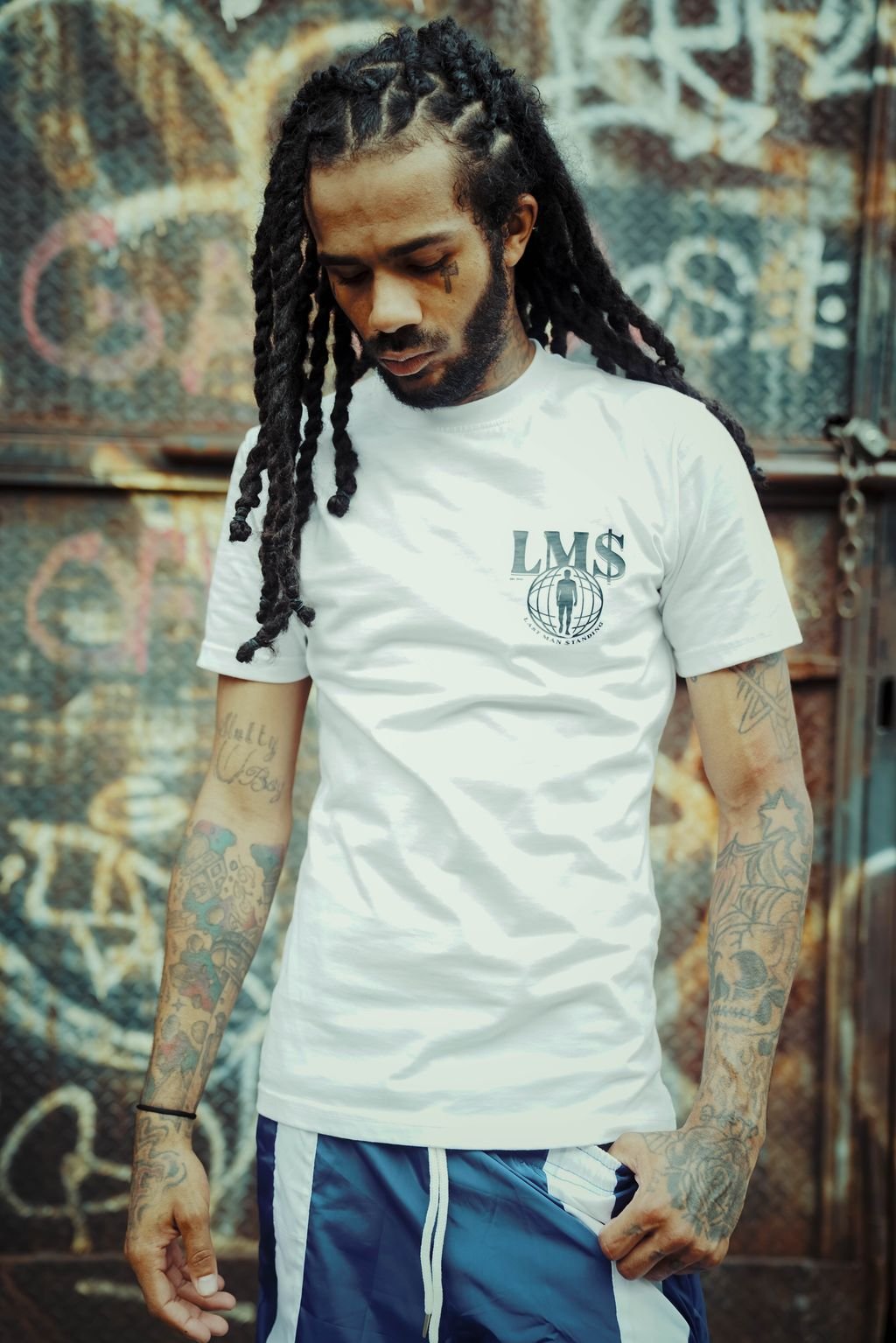 Young man with long dreadlocks and tattoos, wearing a white T-shirt with 'LMS' logo and a globe graphic, standing in front of a graffiti-covered wall.