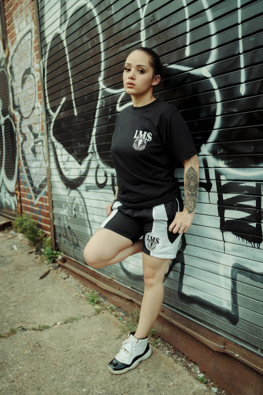 A woman with long dark hair and tattoos on her arm, wearing a black t-shirt and black and white shorts, standing against a graffiti-covered metal garage door.