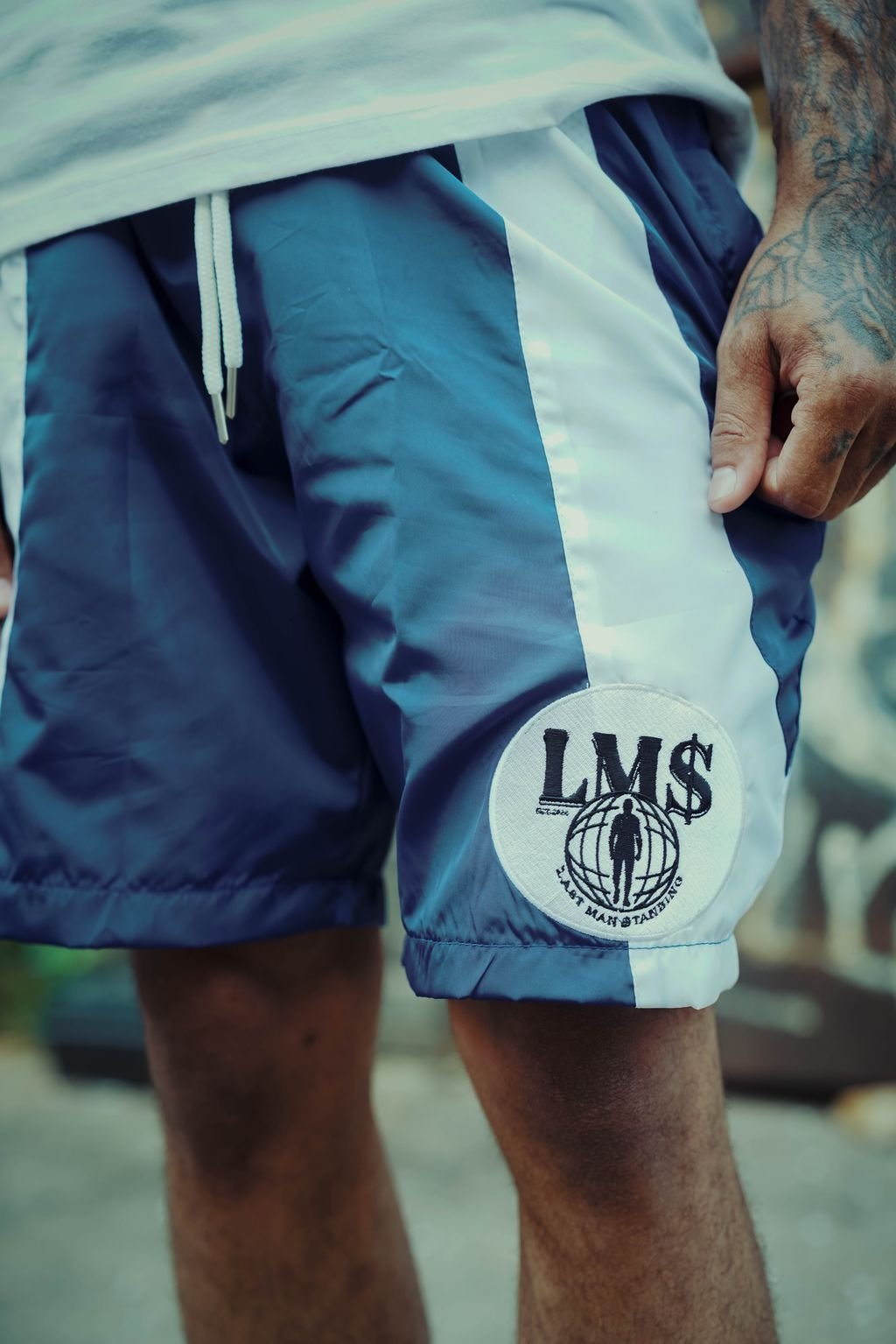 Close-up of a person wearing blue and white shorts with an 'LMS' logo patch on the leg, showing tattooed hands and a portion of a white shirt.