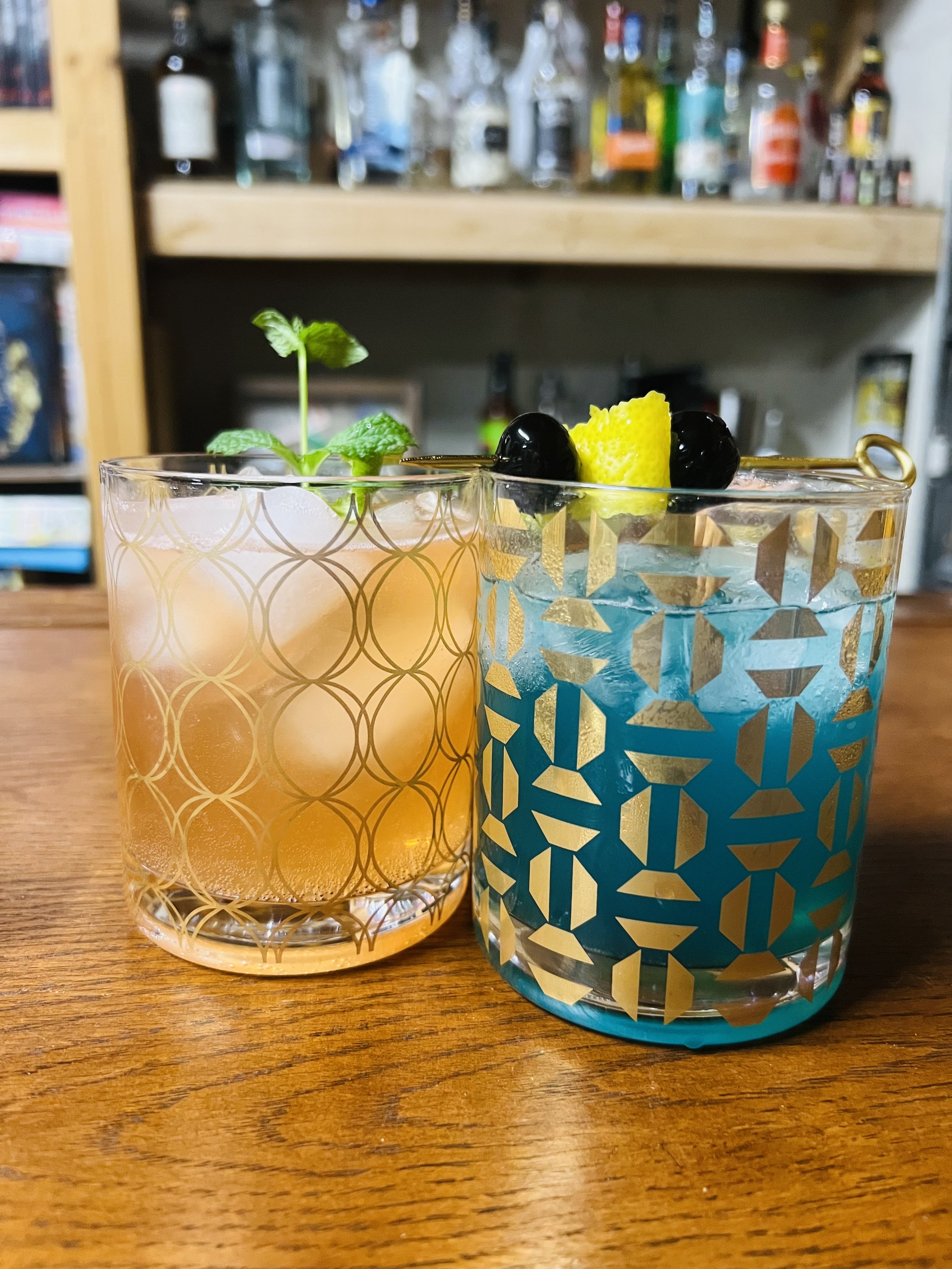 Two colorful cocktails in patterned glasses with garnishes, placed on a wooden table in a bar setting.