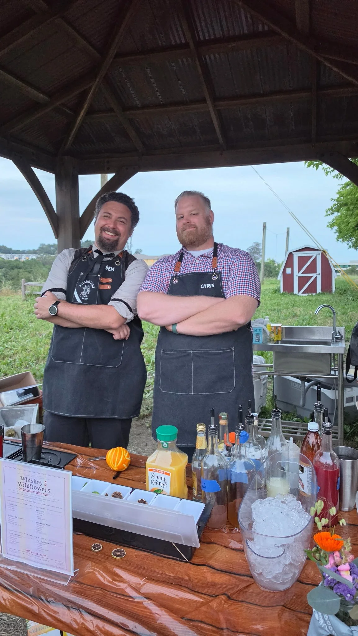 Two men wearing aprons with name tags 'Ben' and 'Chris' stand under a wooden pavilion, smiling with crossed arms, at an outdoor event with a dessert table in front, a grassy background, and a red barn in the distance.
