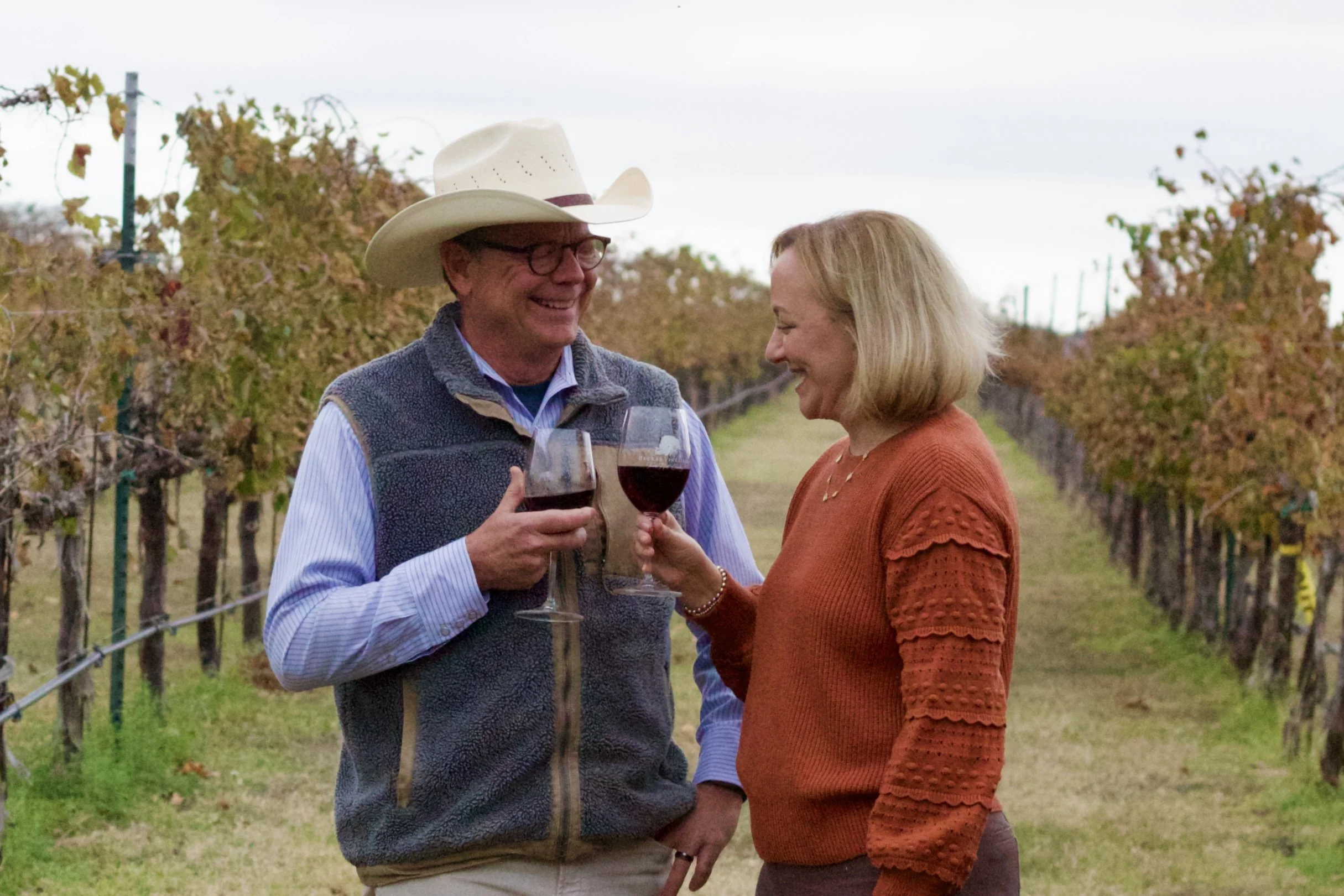 A man and woman in a vineyard holding glasses of red wine, smiling at each other.