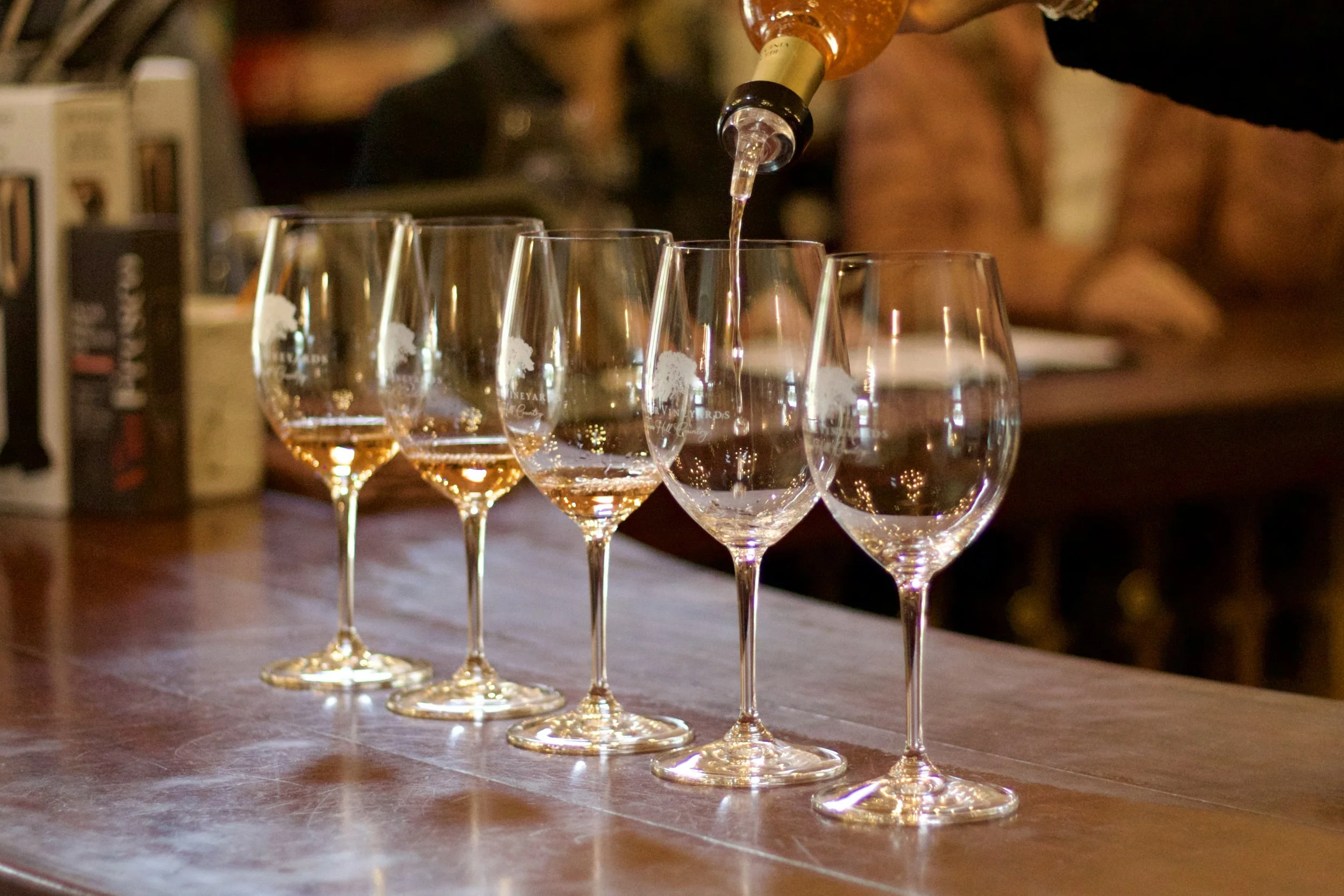 Six champagne glasses are lined up on a wooden bar counter, with golden bubbly liquid being poured into the first glass.