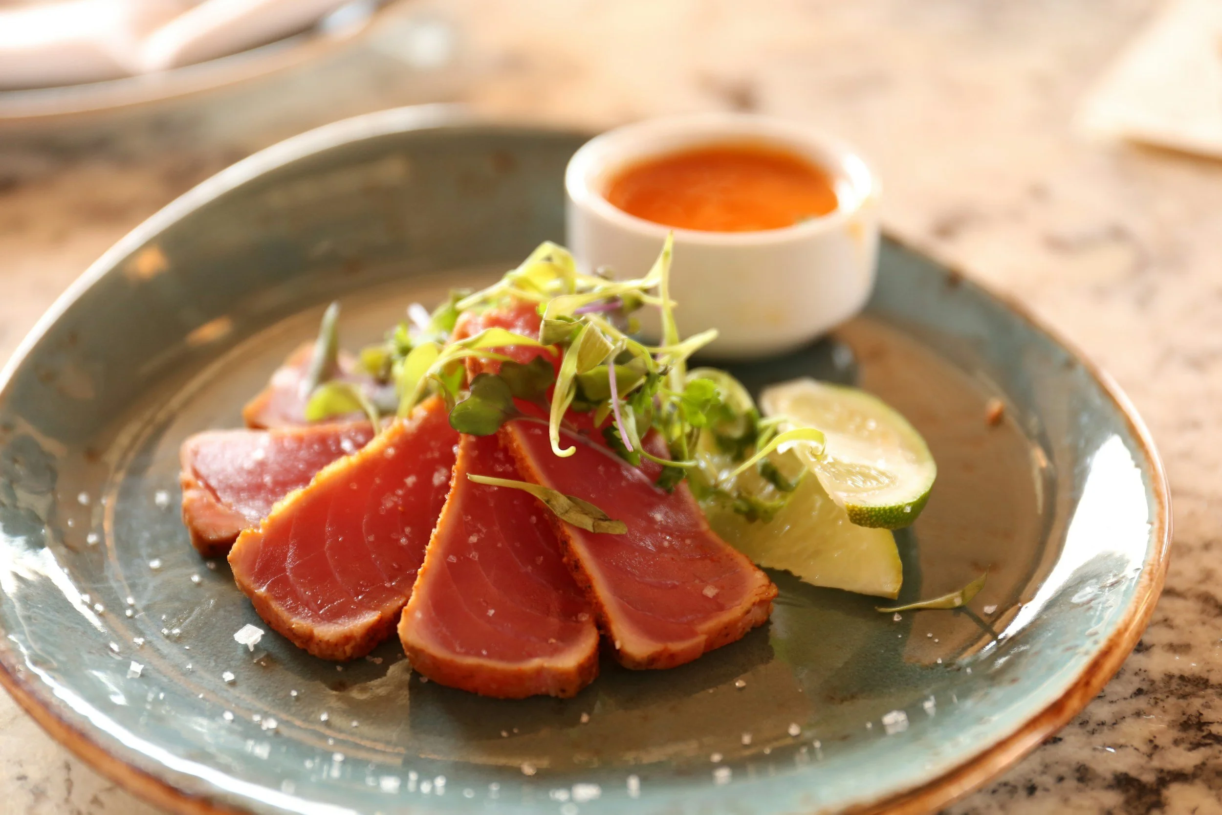 Seared tuna slices garnished with microgreens, lemon and lime wedges, accompanied by a small bowl of dipping sauce on a ceramic plate.