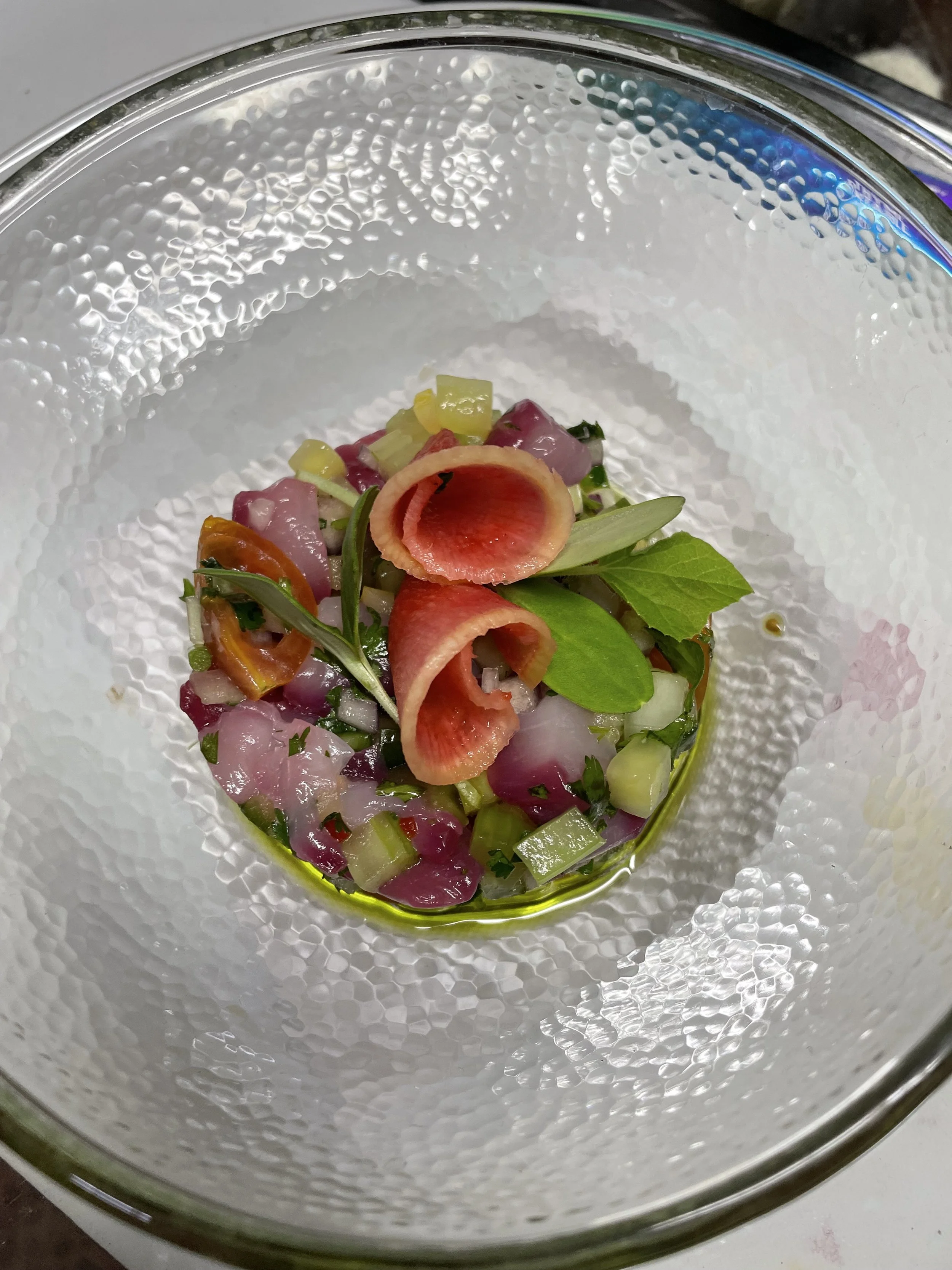 A textured glass bowl containing a colorful salad with chopped vegetables, garnished with fresh herbs and curled vegetable slices.