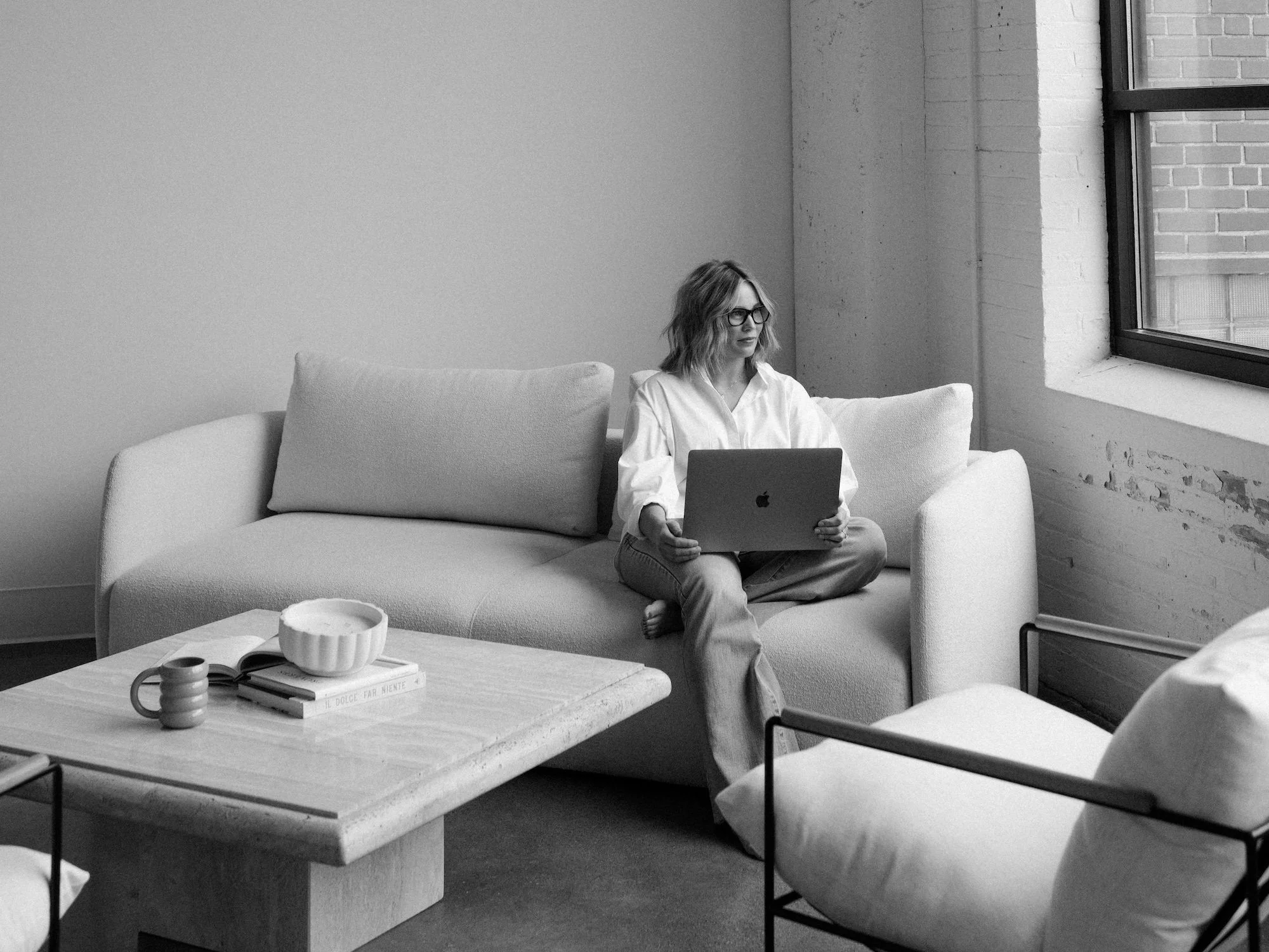 A woman with glasses using a laptop while sitting on a couch in a minimalist living room with a window showing a brick wall outside.