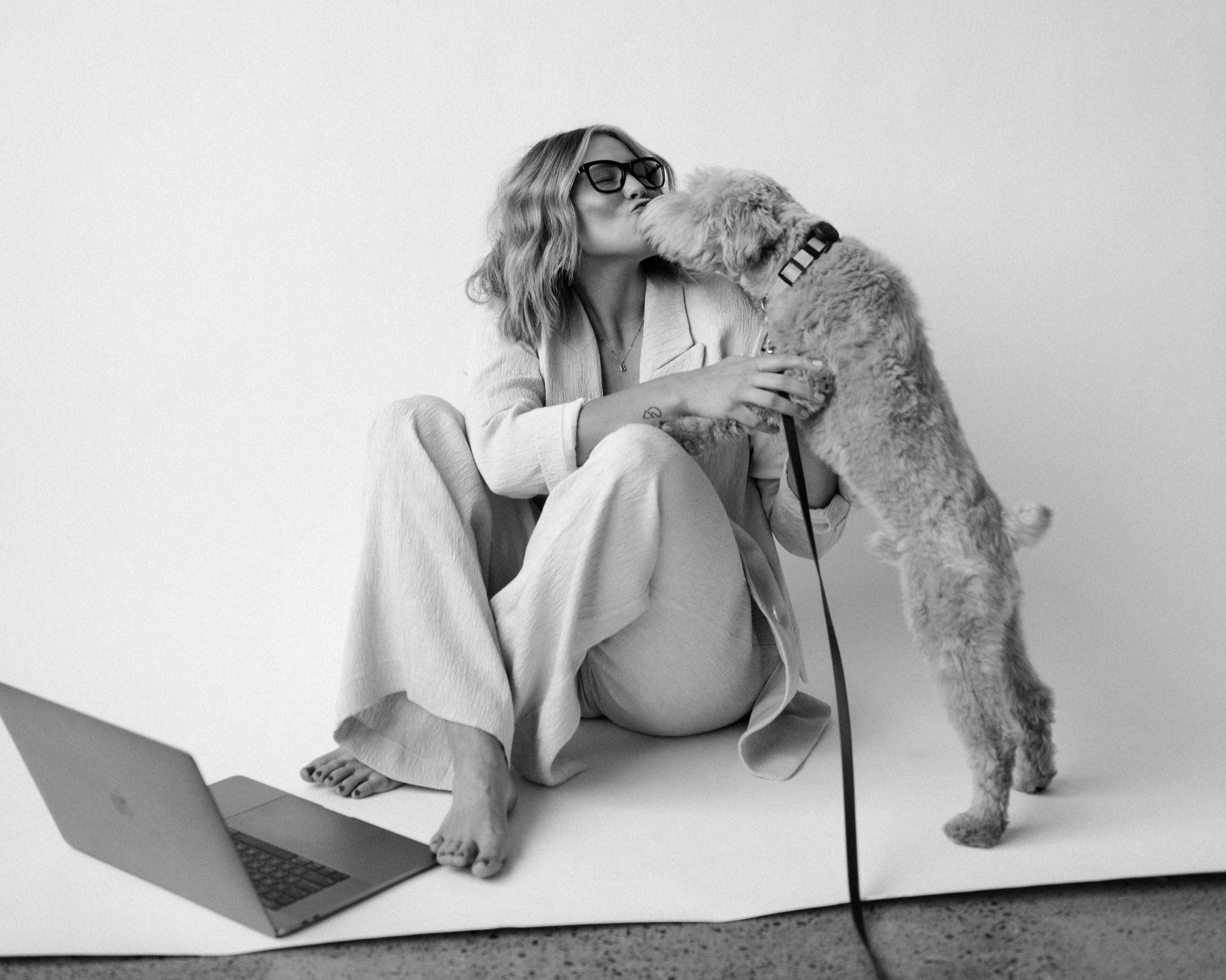 Woman with glasses and wavy hair sitting barefoot on the floor, kissing a small, curly-haired dog, with a laptop on the floor nearby, against a plain background.