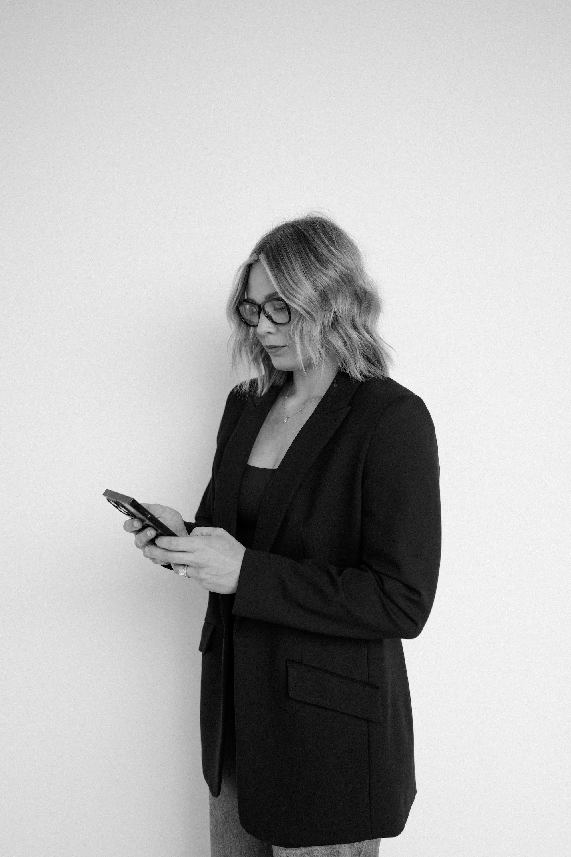 A woman with wavy shoulder-length hair and glasses, wearing a black blazer, is looking at her phone against a plain, light-colored wall.