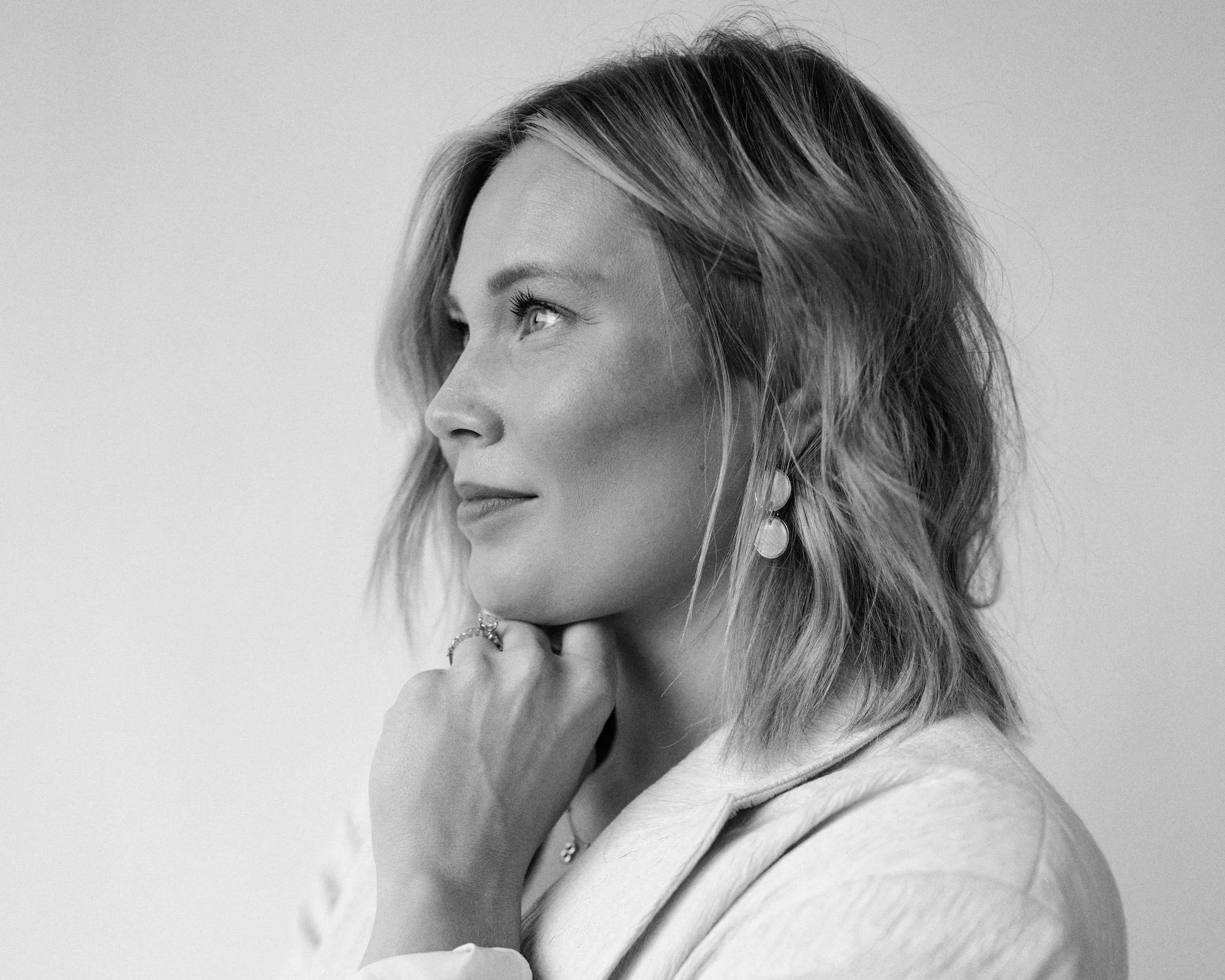 Black and white portrait of a woman with shoulder-length hair, wearing earrings and a ring, looking to the left with her hand resting near her chin.