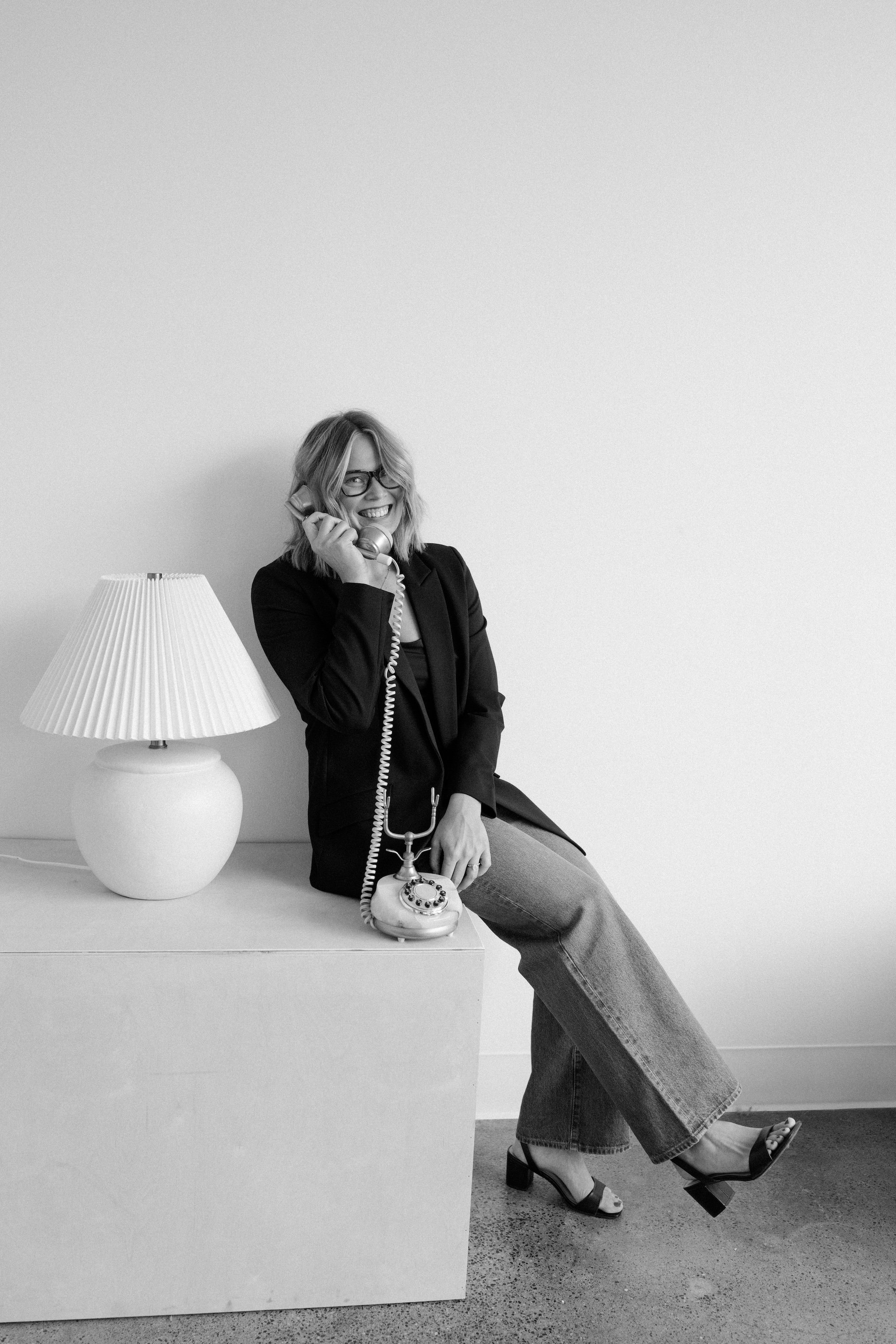 Black and white photo of a woman with glasses, sitting on a rectangular block, talking on a rotary phone, smiling, wearing a blazer, jeans, and heeled sandals, in a room with minimal decor, a lamp on the side.