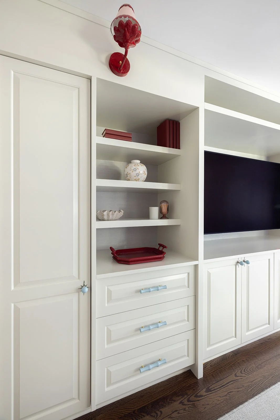 A white built-in entertainment center with open shelves, a large flat-screen TV, and a closed cabinet. The shelves contain decorative items like books, vases, and a red tray, with a red wall-mounted light fixture above.
