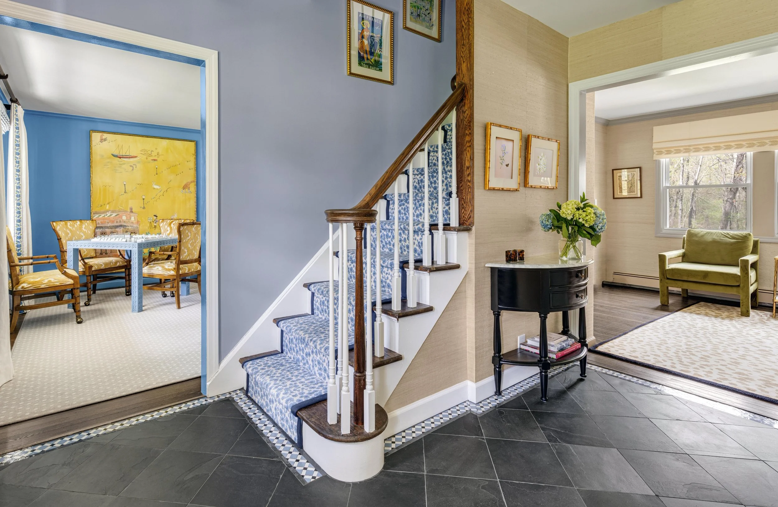 Interior view of a staircase with blue patterned carpet, leading to a dining room with yellow chairs and artwork on the walls, and a living room with a green chair, large window, and floral arrangements.