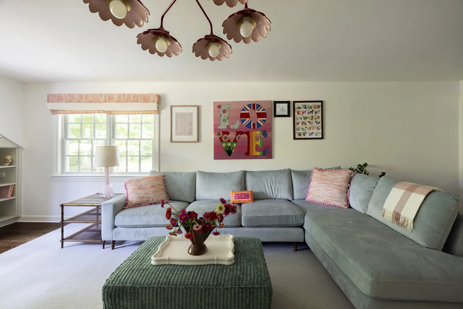 Cozy living room with a light gray sectional sofa, pink pillows, a green textured coffee table with a white tray holding a vase of pink and red flowers, a window with a valance, wall art that spells LOVE with colorful letters, and a white bookshelf w