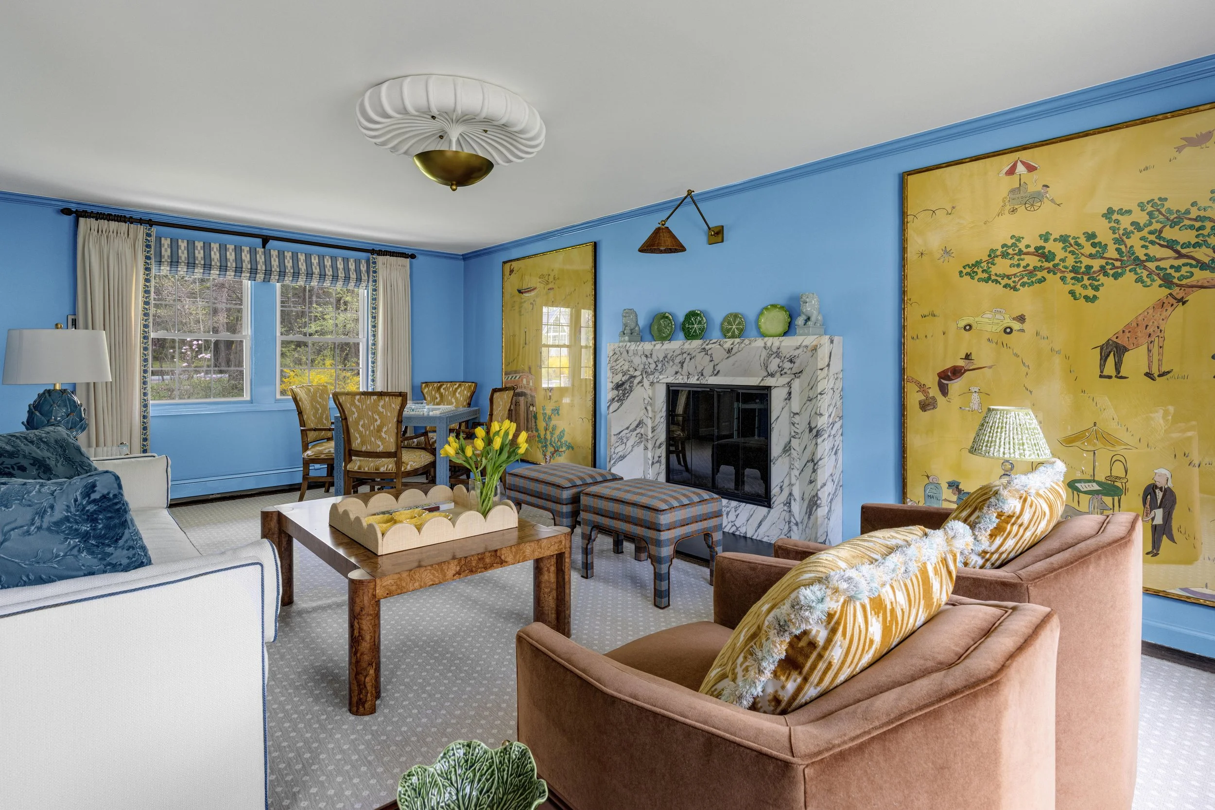 Living room with blue walls, large windows with white curtains, a marble fireplace, yellow and blue wallpaper, and furniture including a white sofa, brown armchair, and yellow-patterned pillows, with a vase of yellow tulips on a wooden coffee table.