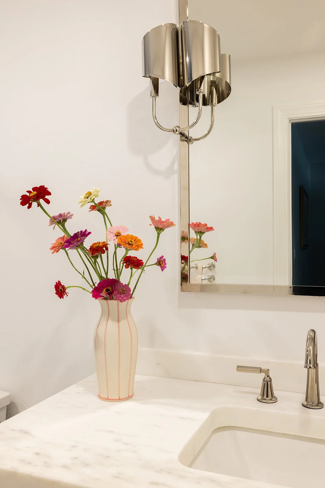 A white bathroom countertop with a vase of colorful flowers, a mirror, a wall-mounted light fixture, a sink with a faucet, and a partially visible mirror reflected on the wall.