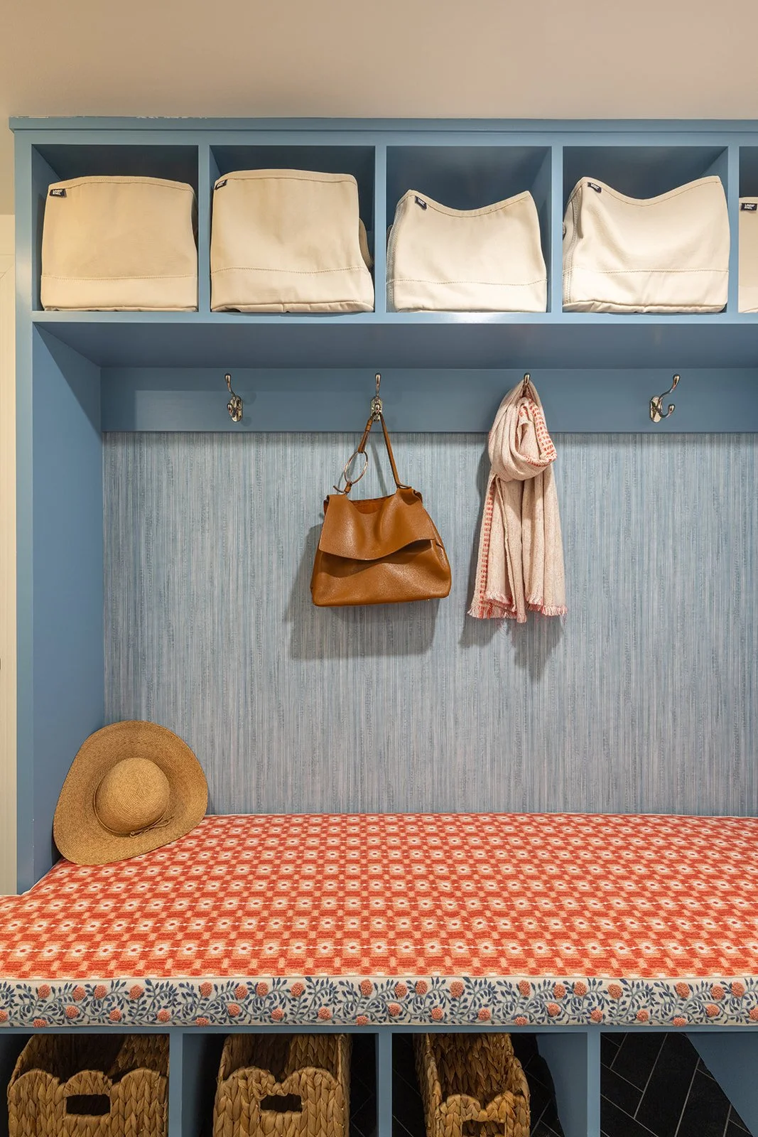Blue cubby storage unit with white fabric bins on top, hooks with a brown purse and a pink scarf hanging, a straw hat on a red patterned bench, and woven baskets underneath.